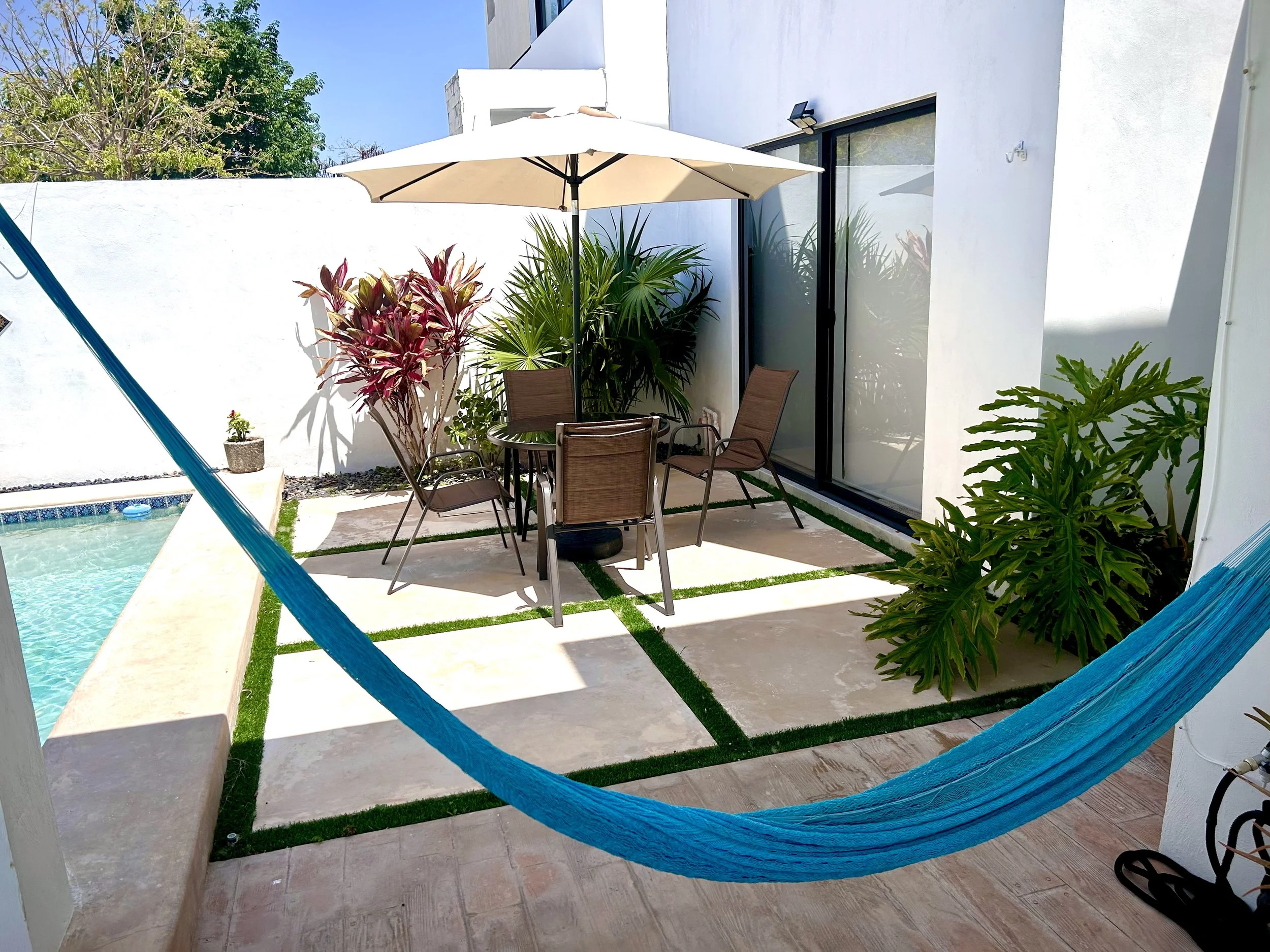 A backyard patio with a swimming pool on the left, a round table with chairs and a large umbrella in the middle, and tropical plants against a white wall. A blue hammock is in the foreground.
