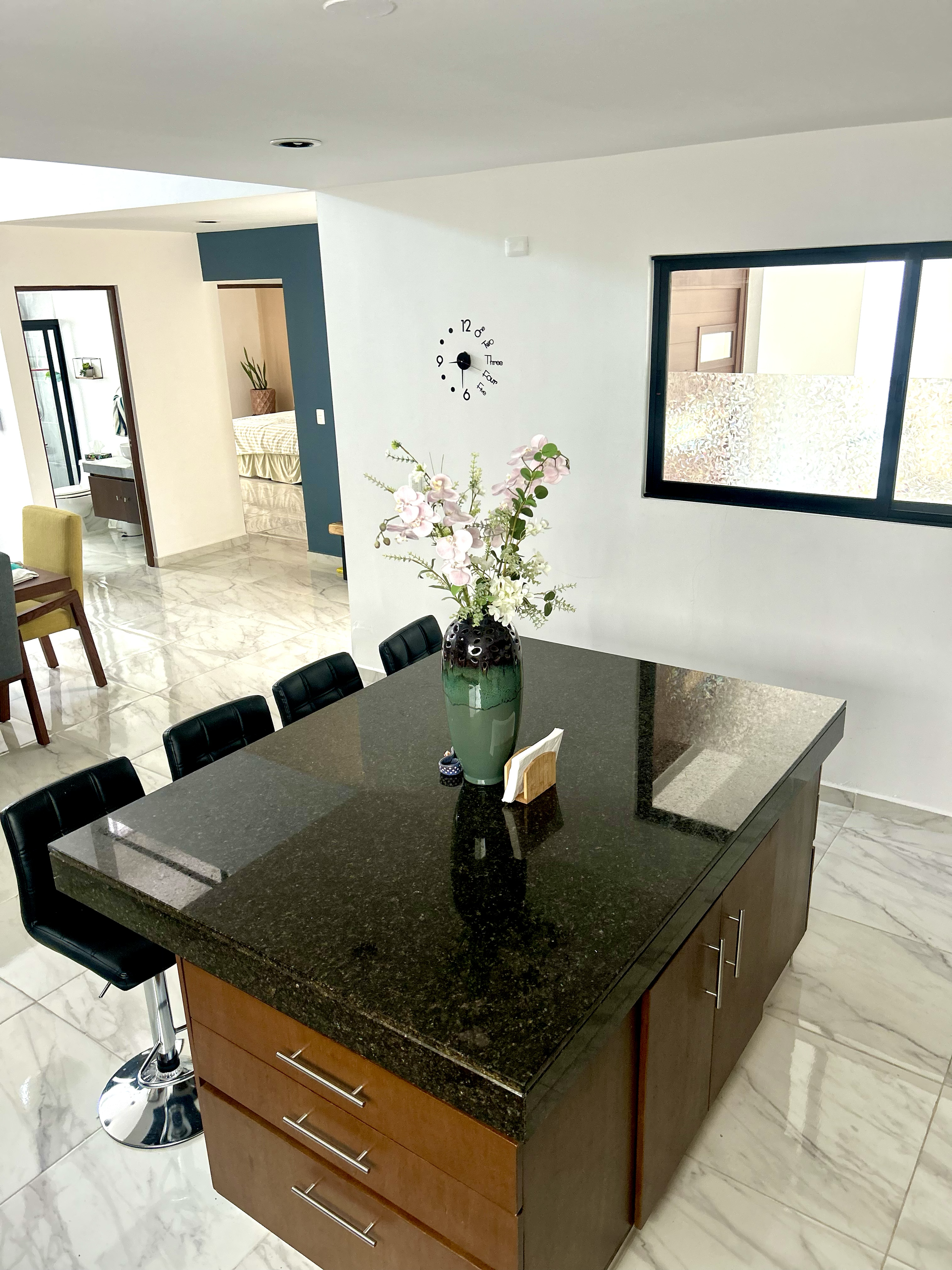 Modern kitchen with a black granite island, a green and black vase with white flowers, and black bar stools. White marble floors, a white wall with a black clock, and large windows.