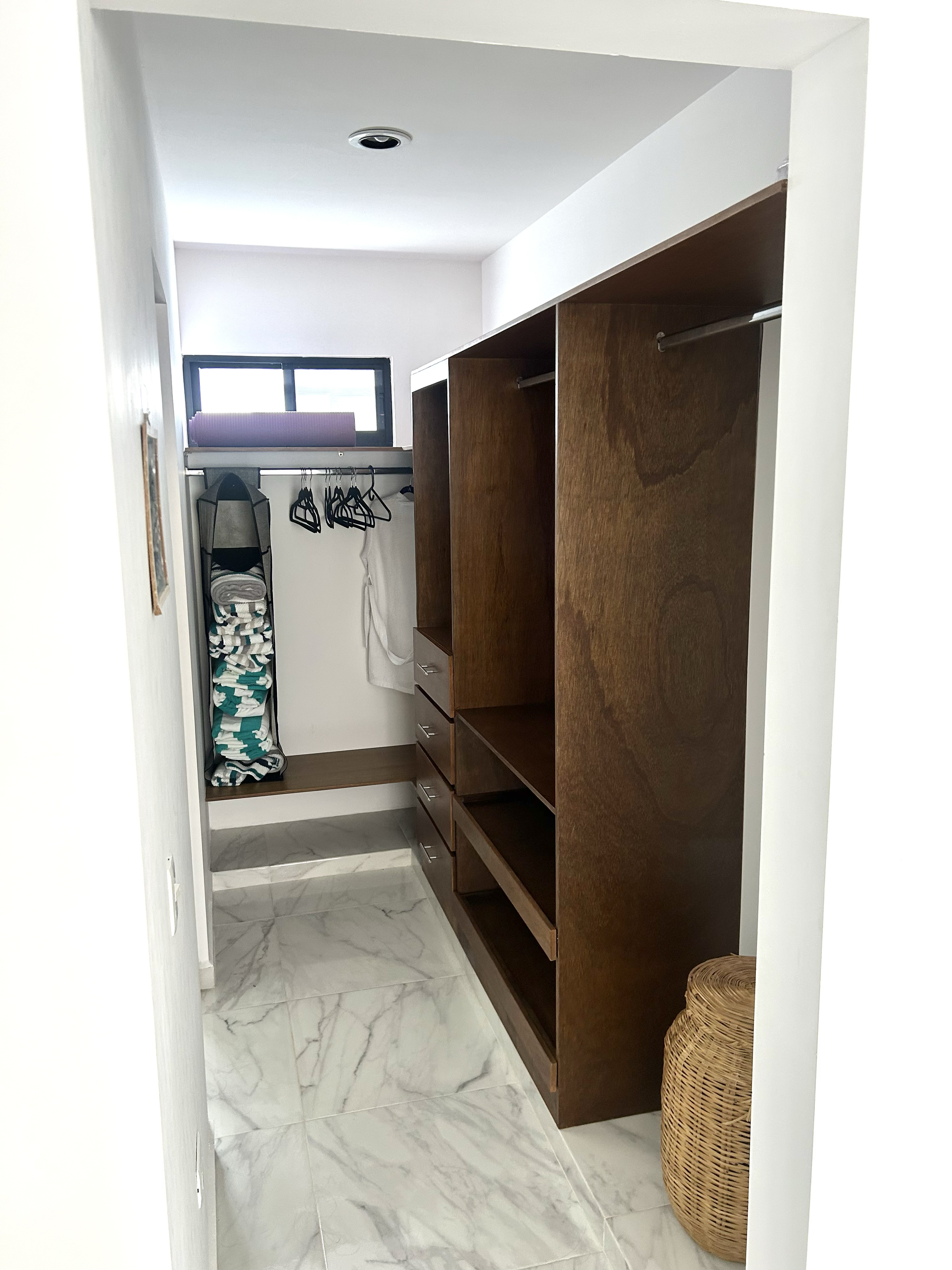A walk-in closet with white walls, marble floor, and a window. It contains wooden shelving, drawers, and hanging rods, with some folded clothes and hangers. A wicker basket is on the right side.