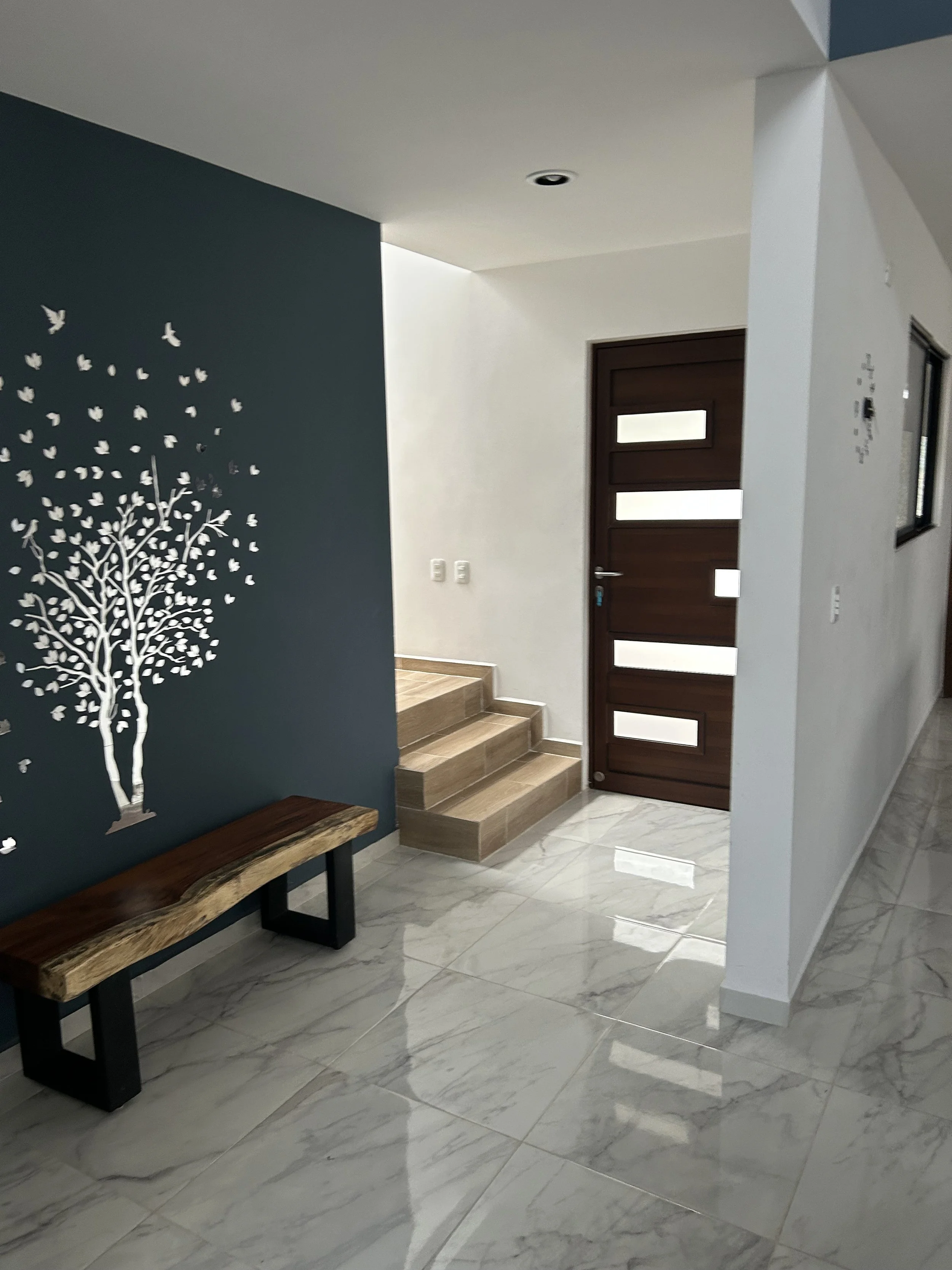 Interior view of a modern home with a wooden front door featuring horizontal glass panels, a marble tiled floor, a blue accent wall with a white tree and bird decal, and a small wooden bench.