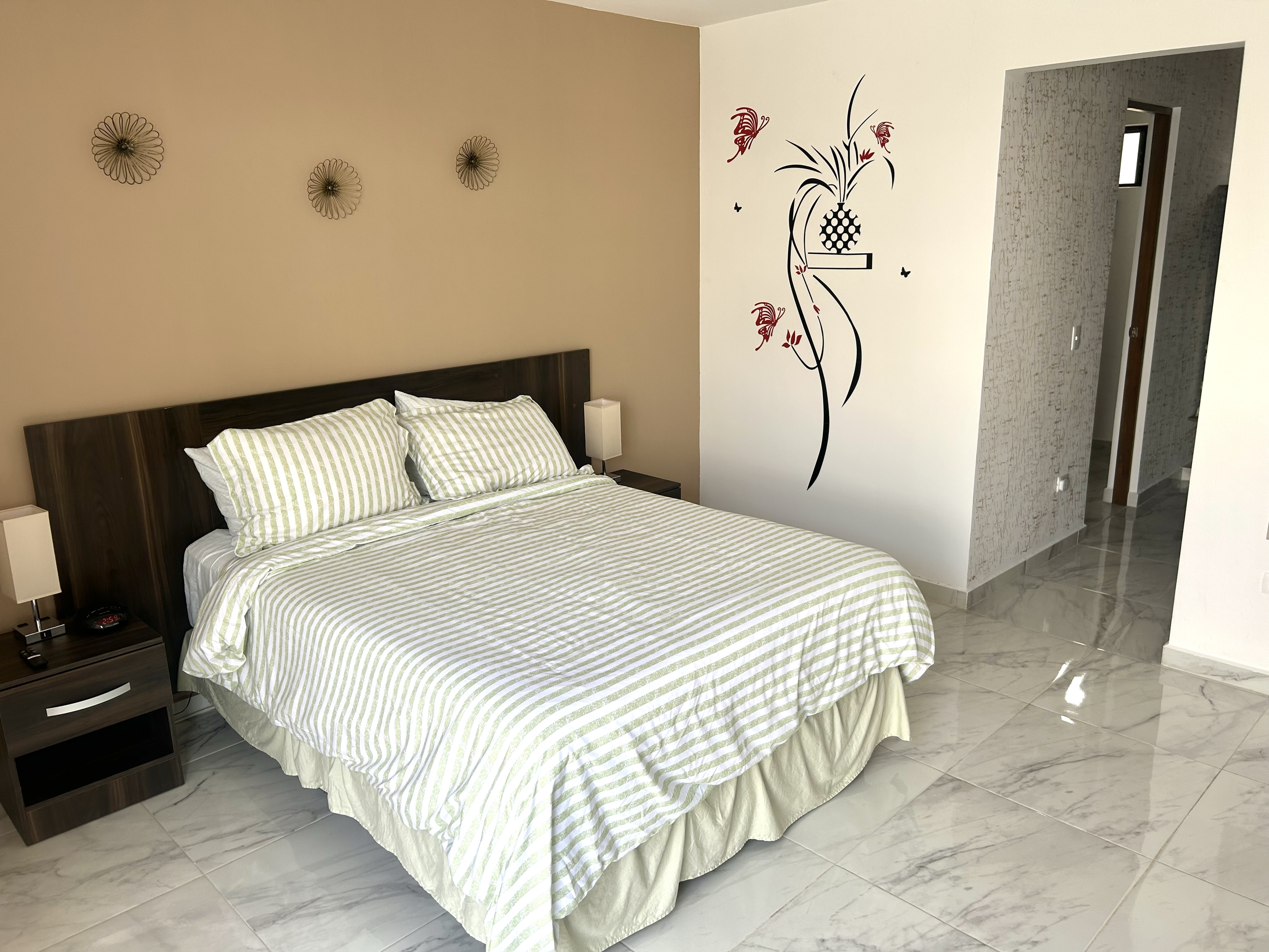A bedroom with a bed, striped green and white bedding, wooden headboard, two bedside tables with lamps, beige wall with black metal decorative flowers, and a white wall with a red and black floral mural, inside a room with marble flooring.