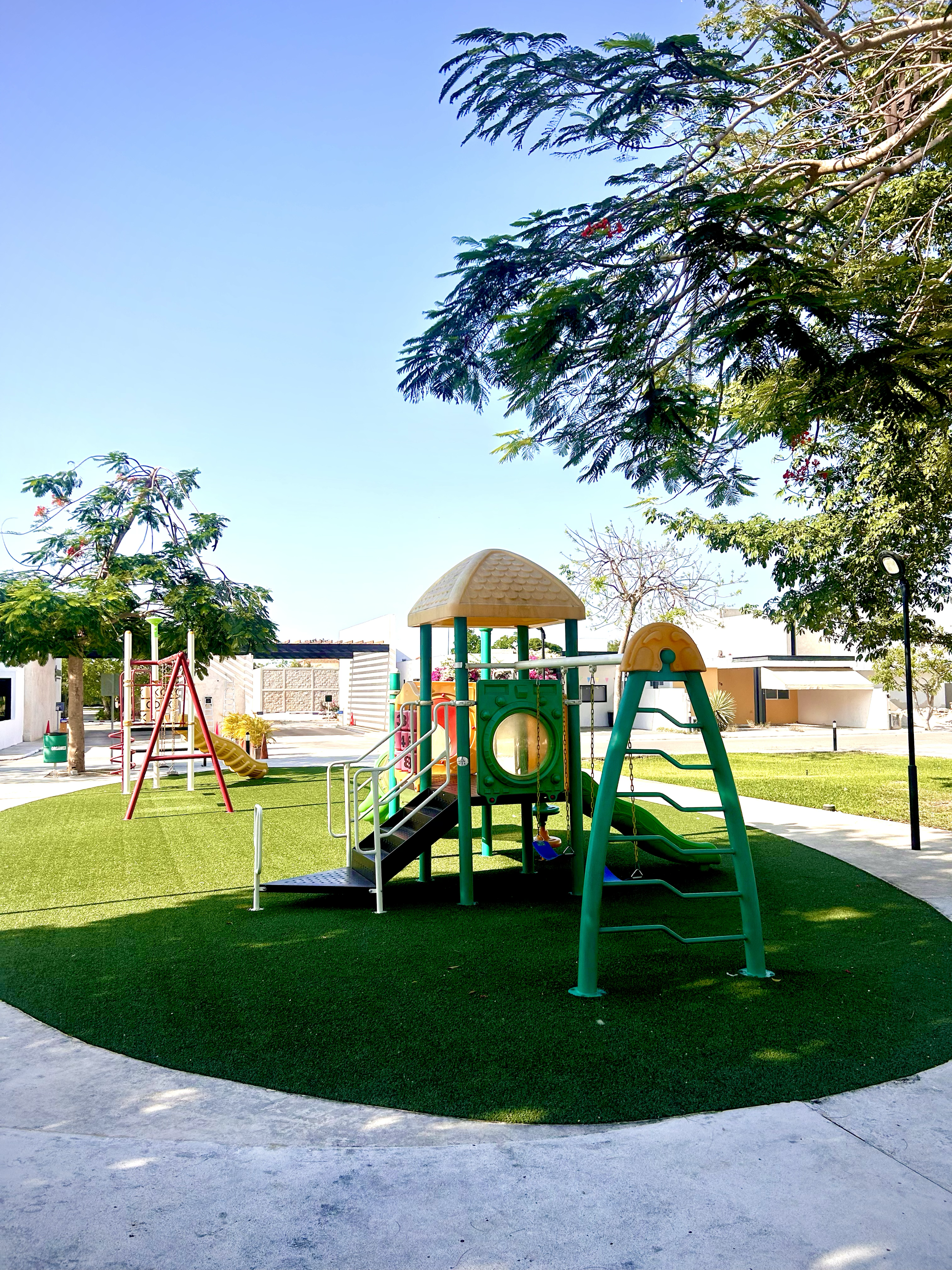 Colorful playground with slides, swings, and climbing structures, surrounded by green grass and trees on a sunny day.