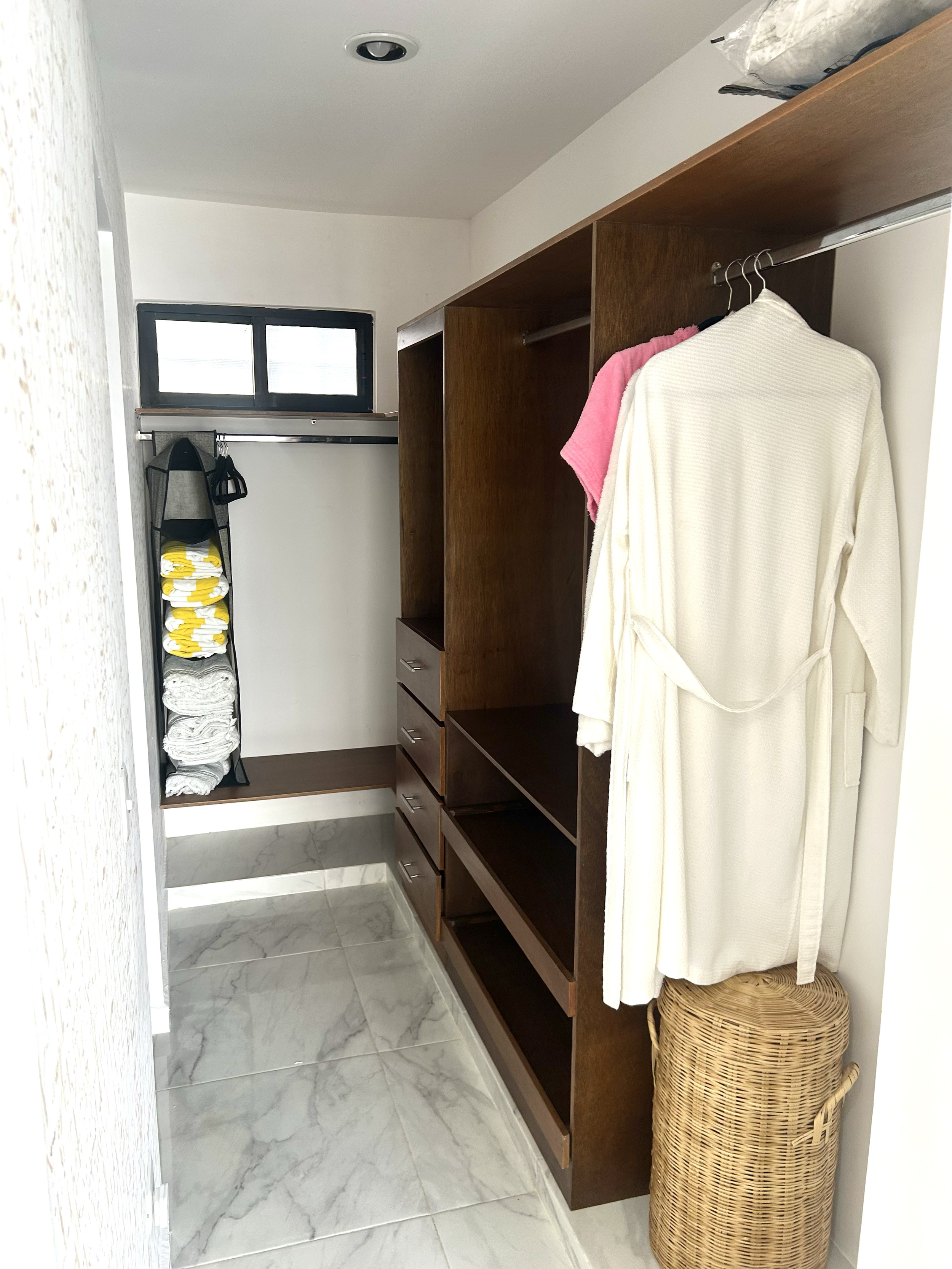 Empty walk-in closet with wooden shelving, hanging rail with clothing, a laundry basket, and folded towels on a vertical rack, with a small window at the end.
