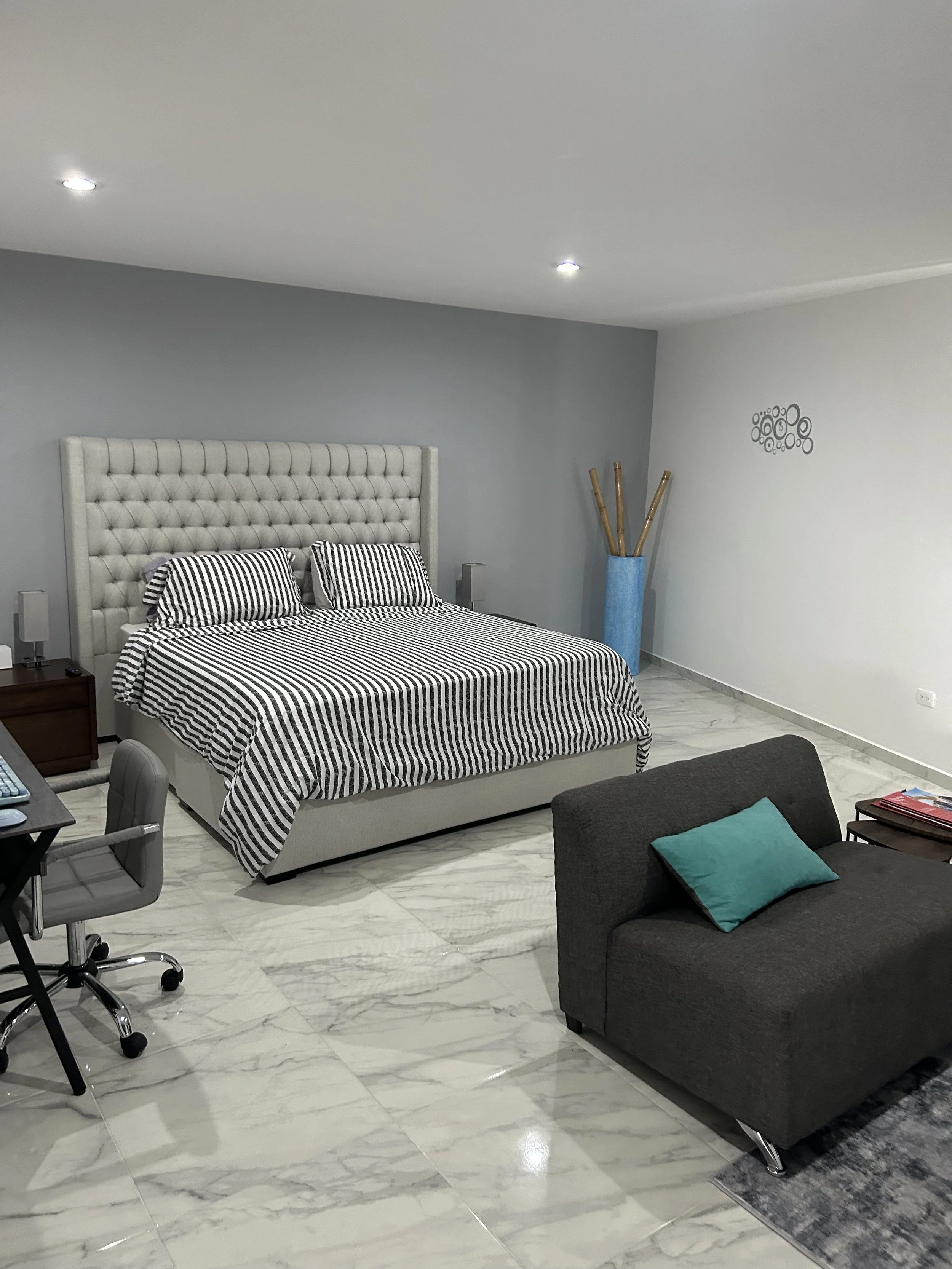 Modern bedroom with a large bed, black and white striped bedding, a gray upholstered headboard, a gray armchair with teal pillow, a small wooden table, a black desk with gray chair, and a blue vase with bamboo sticks, marble floor, and white ceiling 