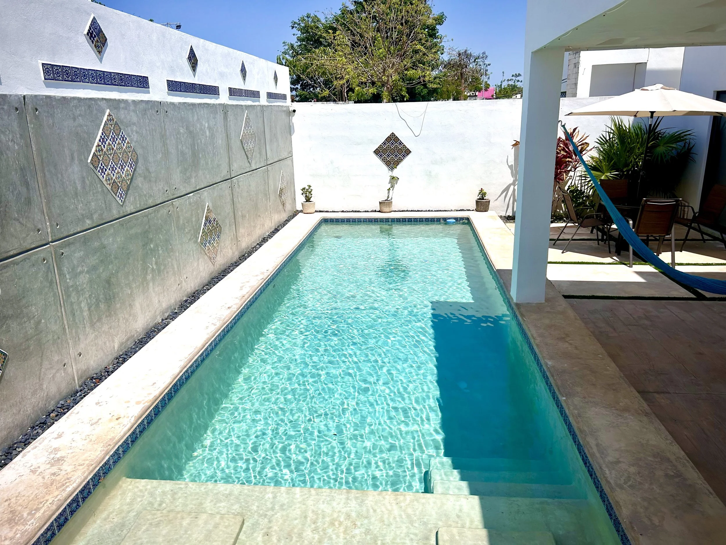 A backyard swimming pool with clear water, surrounded by a concrete border and a white wall with decorative tiles. To the right, there is a patio area with a white umbrella, potted plants, and outdoor furniture, including a hammock.