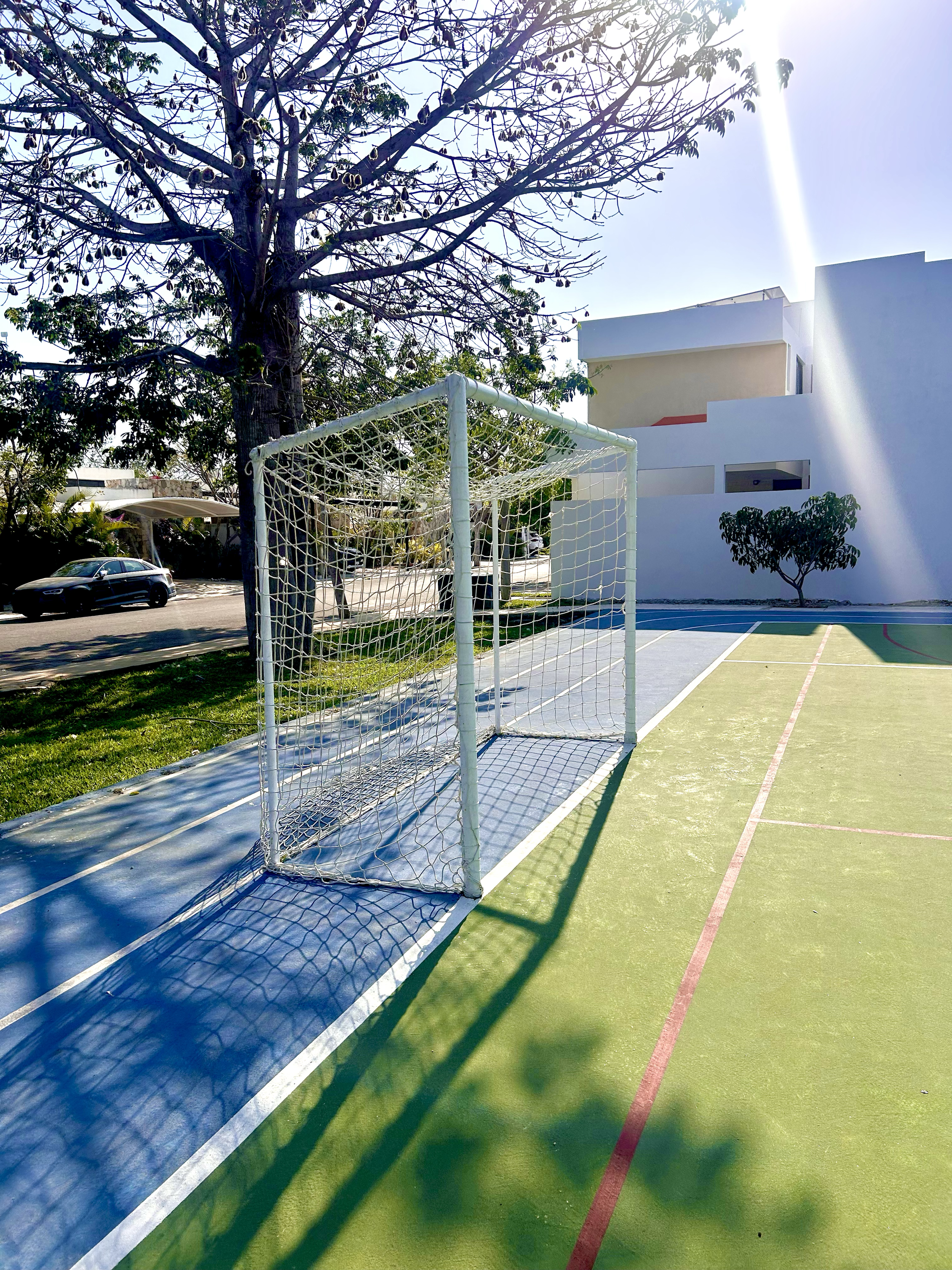 An outdoor futsal court with a white goalpost. There are trees, parked cars, a modern white building, and a clear sky with sunlight and shadows.