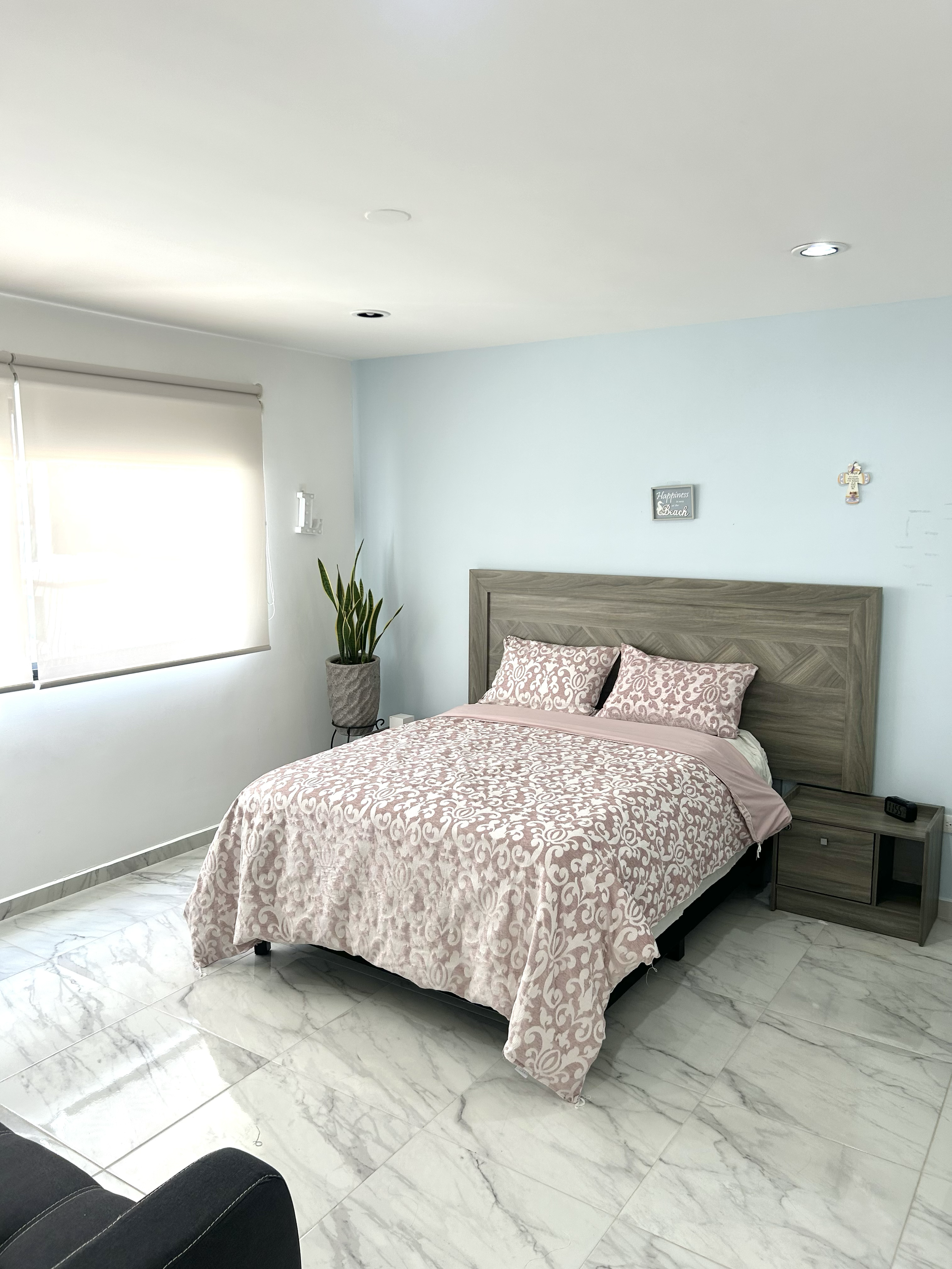 A bedroom with a bed featuring pink and white patterned bedding, a wooden headboard, and pillows, next to a window with closed blinds, a potted plant, and a nightstand with a clock, on a marble floor.