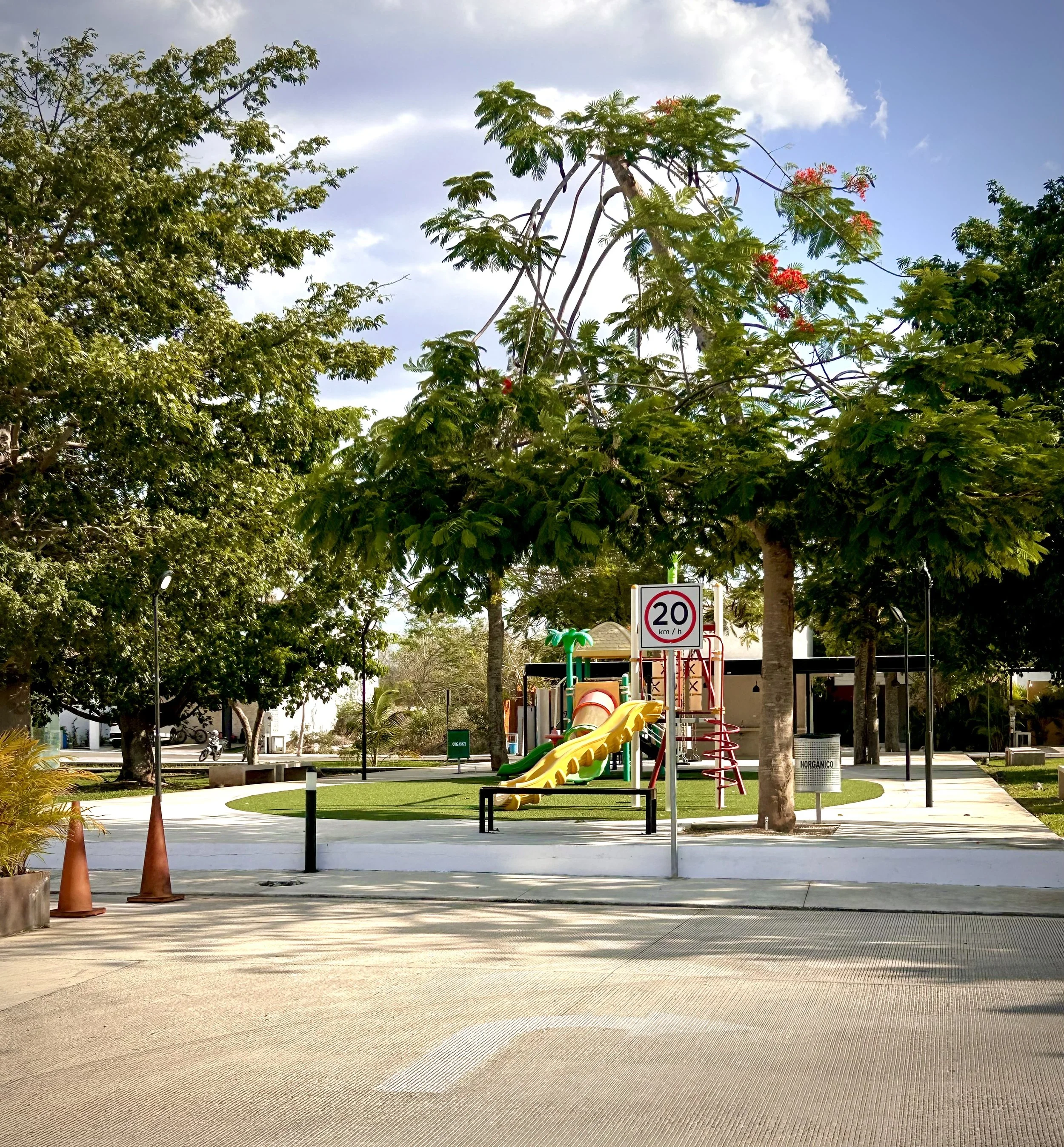 A playground in a park with colorful slides and climbing structures, surrounded by trees and a sidewalk. There are cones and a speed limit sign of 20 km/h in the foreground.