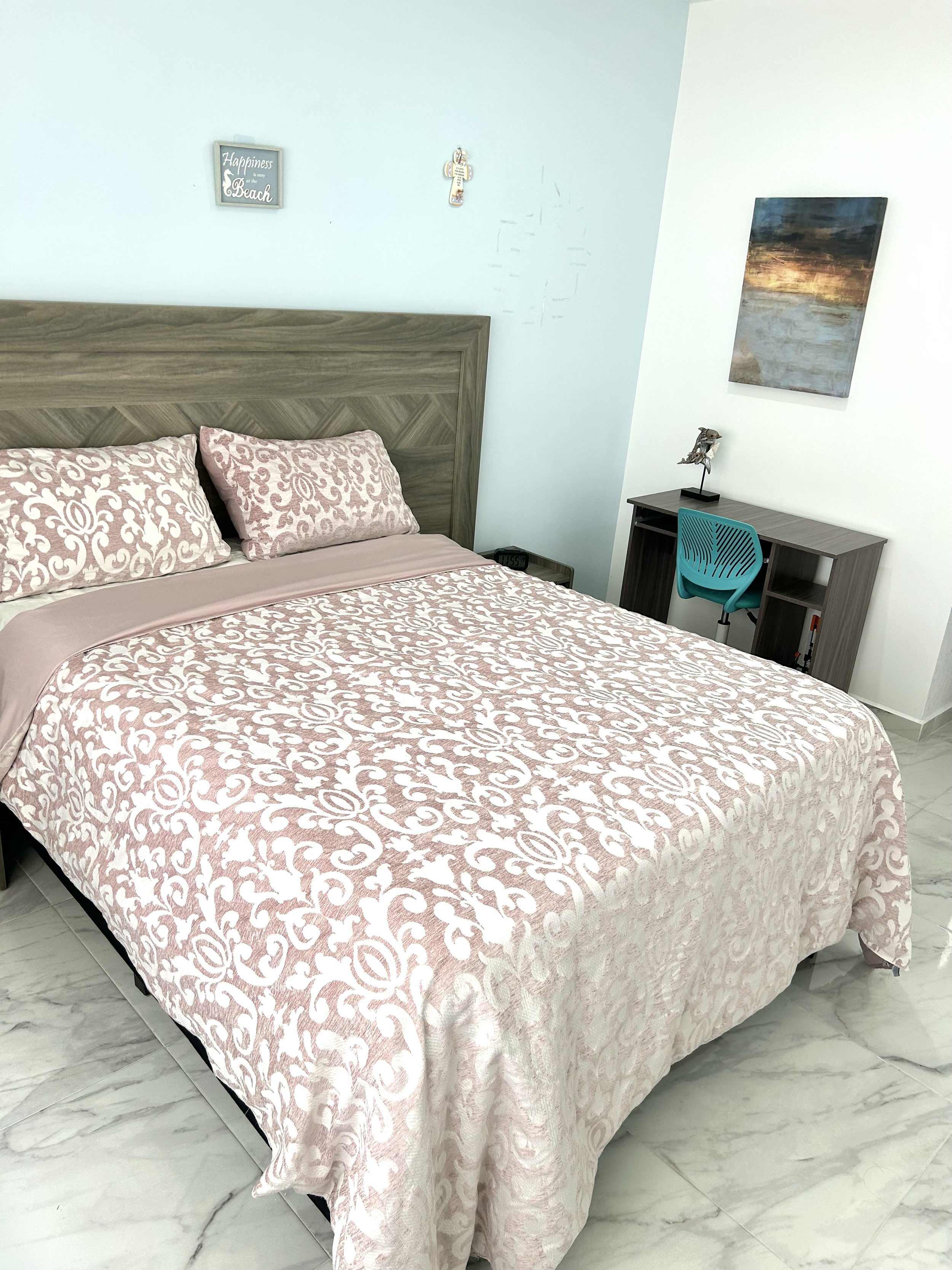 A bedroom with a queen-sized bed covered in a pink and white patterned bedspread and matching pillows. There is a wooden headboard, a small desk with a blue chair, wall art, and a clock on the side table. The floor is tiled in white and gray.