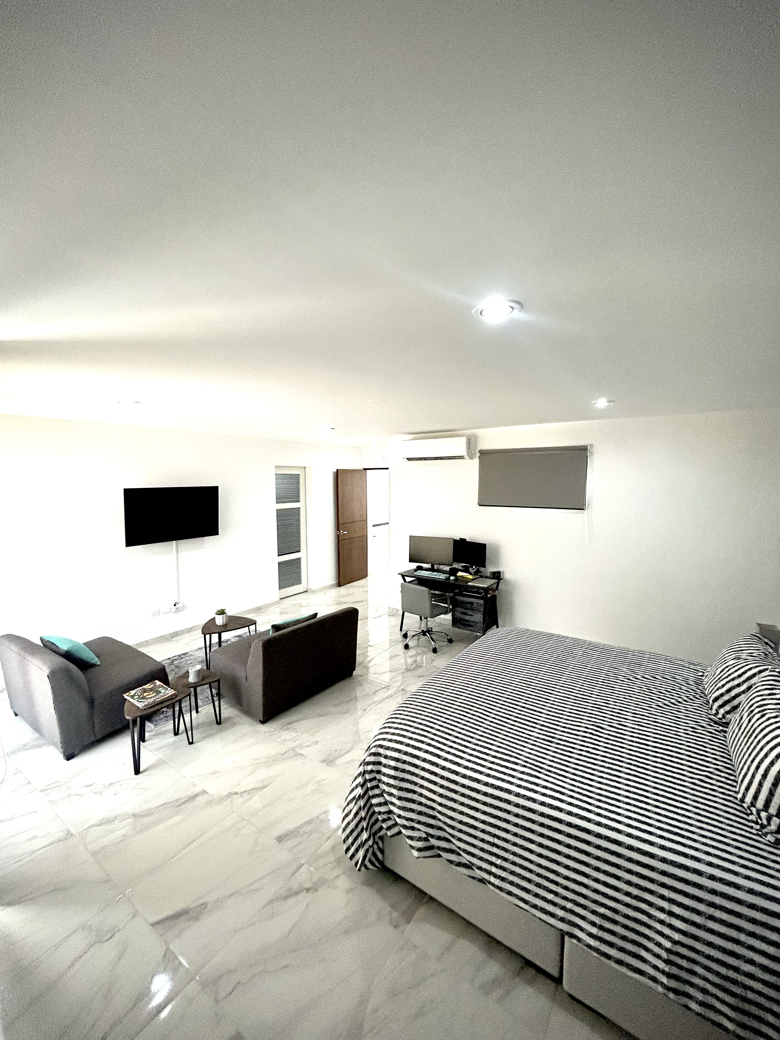 A hotel room with a bed covered in black and white striped bedding, a sitting area with two sofas and a small table, a wall-mounted TV, a desk with a computer, and a window with a roller blind.