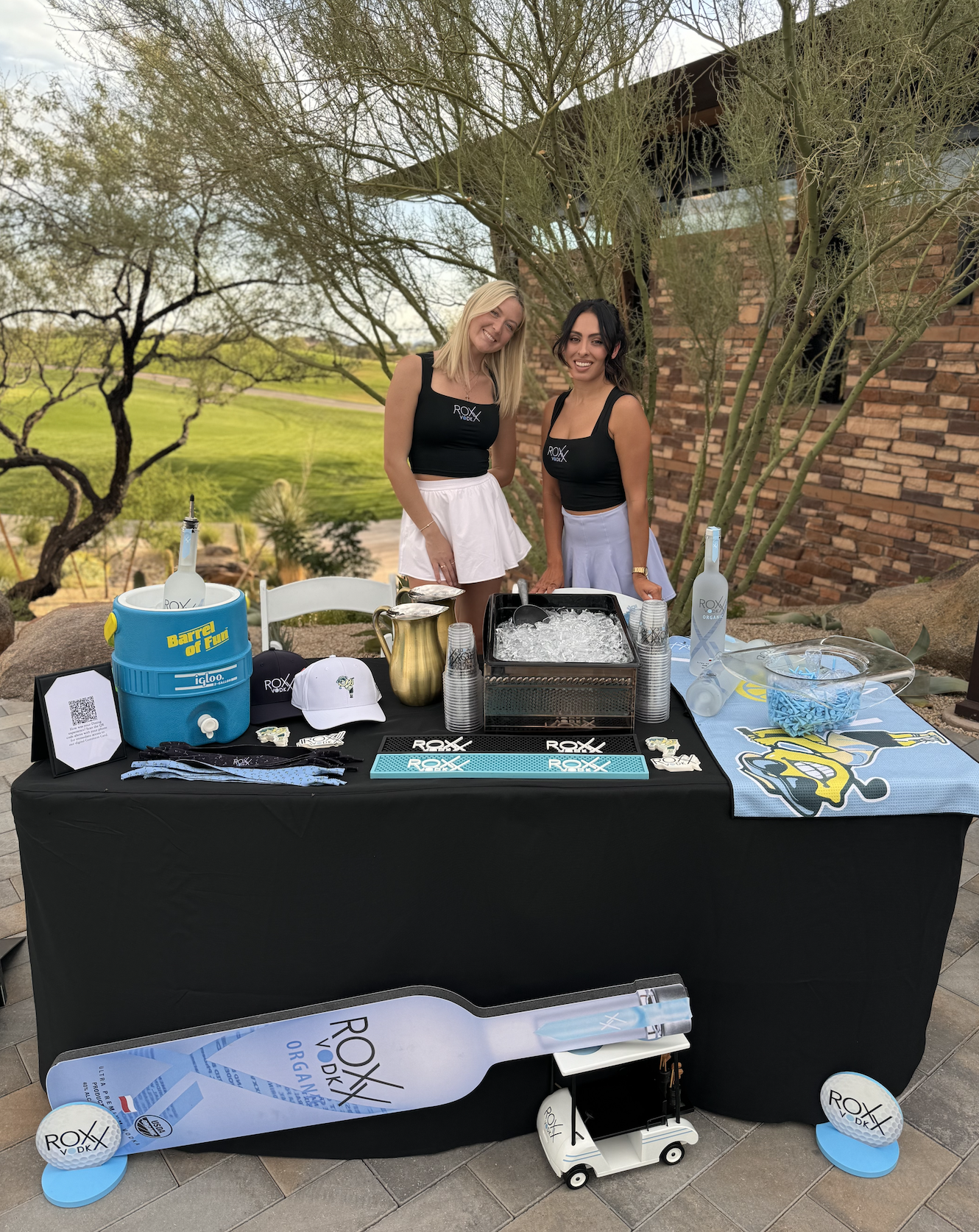 Two women standing behind a table at an outdoor event, with various promotional items, drinks, and a large bottle-shaped sign on the table, with trees and a brick building in the background.
