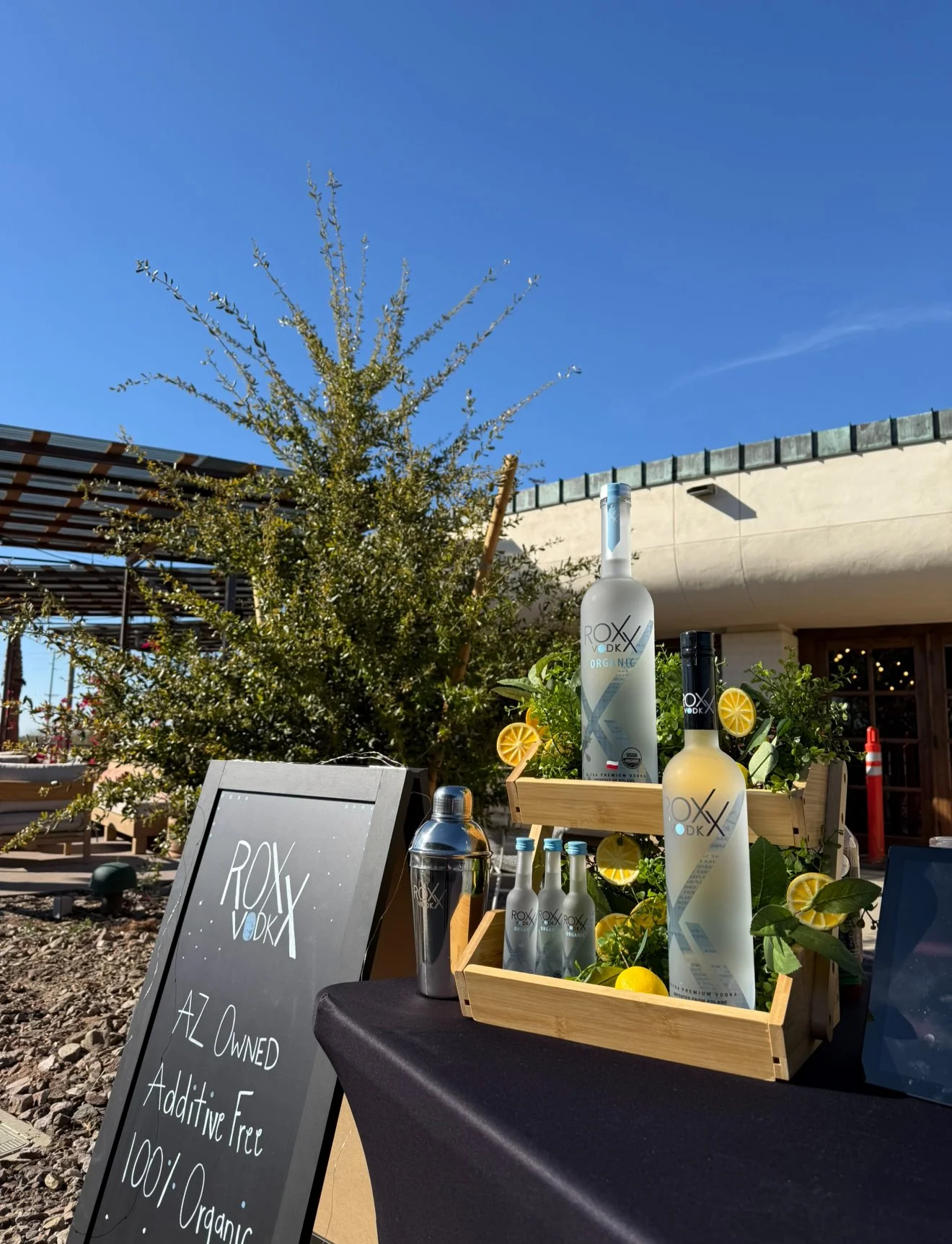 Display of Roxy Vodka bottles and a blackboard sign at an outdoor event on a sunny day.