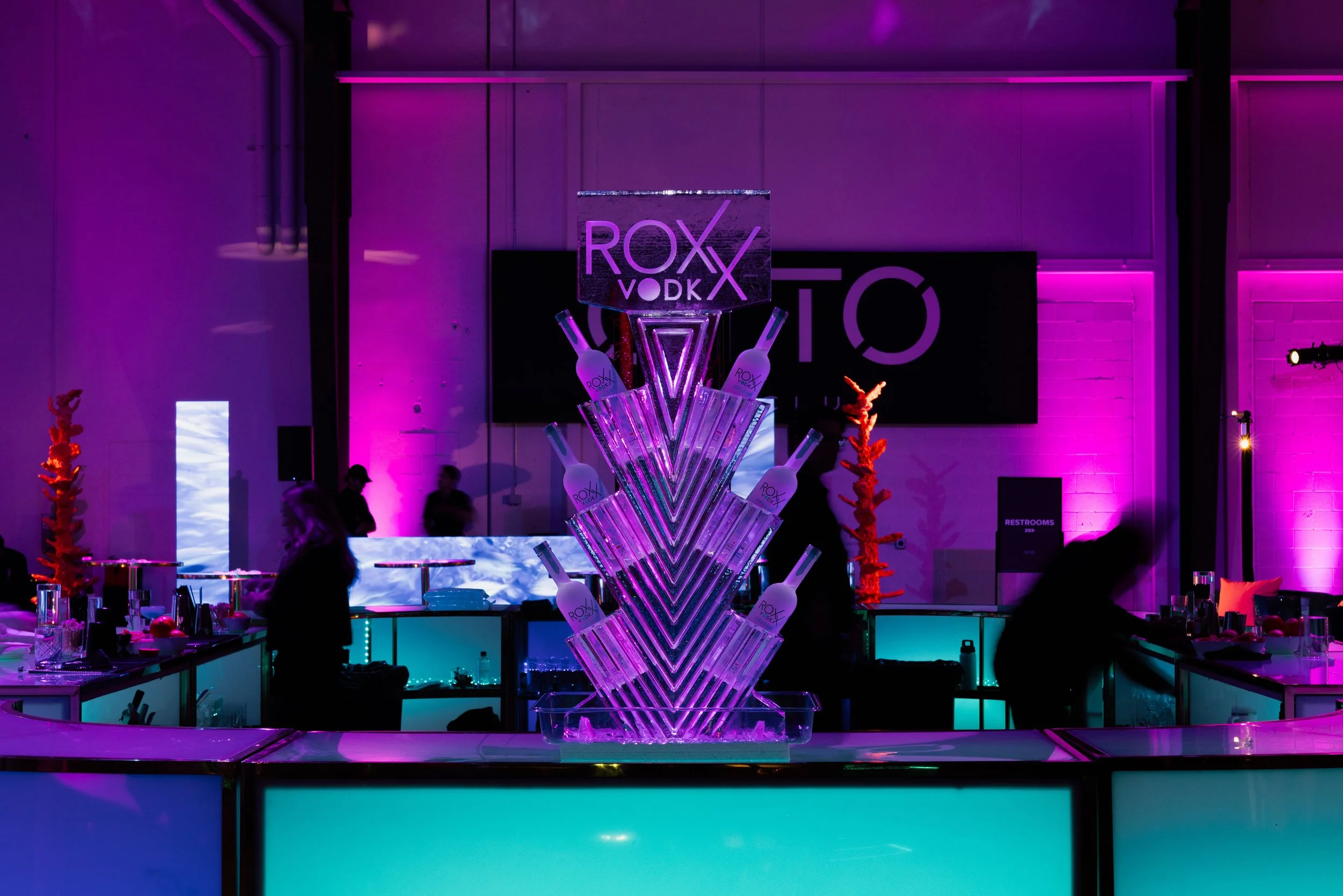 A LED-lit display with bottles of Roxx Vodka arranged in a geometric, tower-like structure at a vibrant event space illuminated with purple and teal lighting.