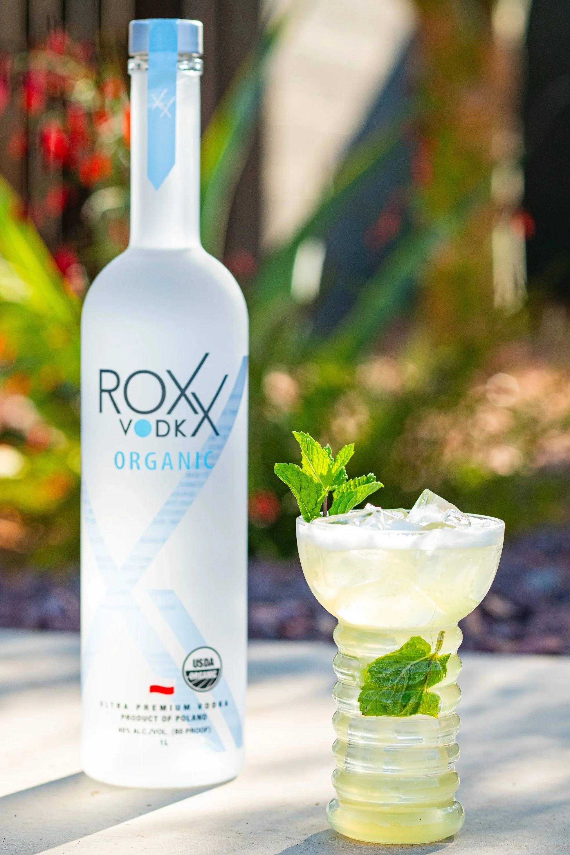 A white bottle of Roxi Vodka Organic with a blue cap and label standing next to a glass filled with a light-colored beverage, ice, and garnished with fresh mint leaves on a white surface outdoors, with a blurred background of greenery and red berries.