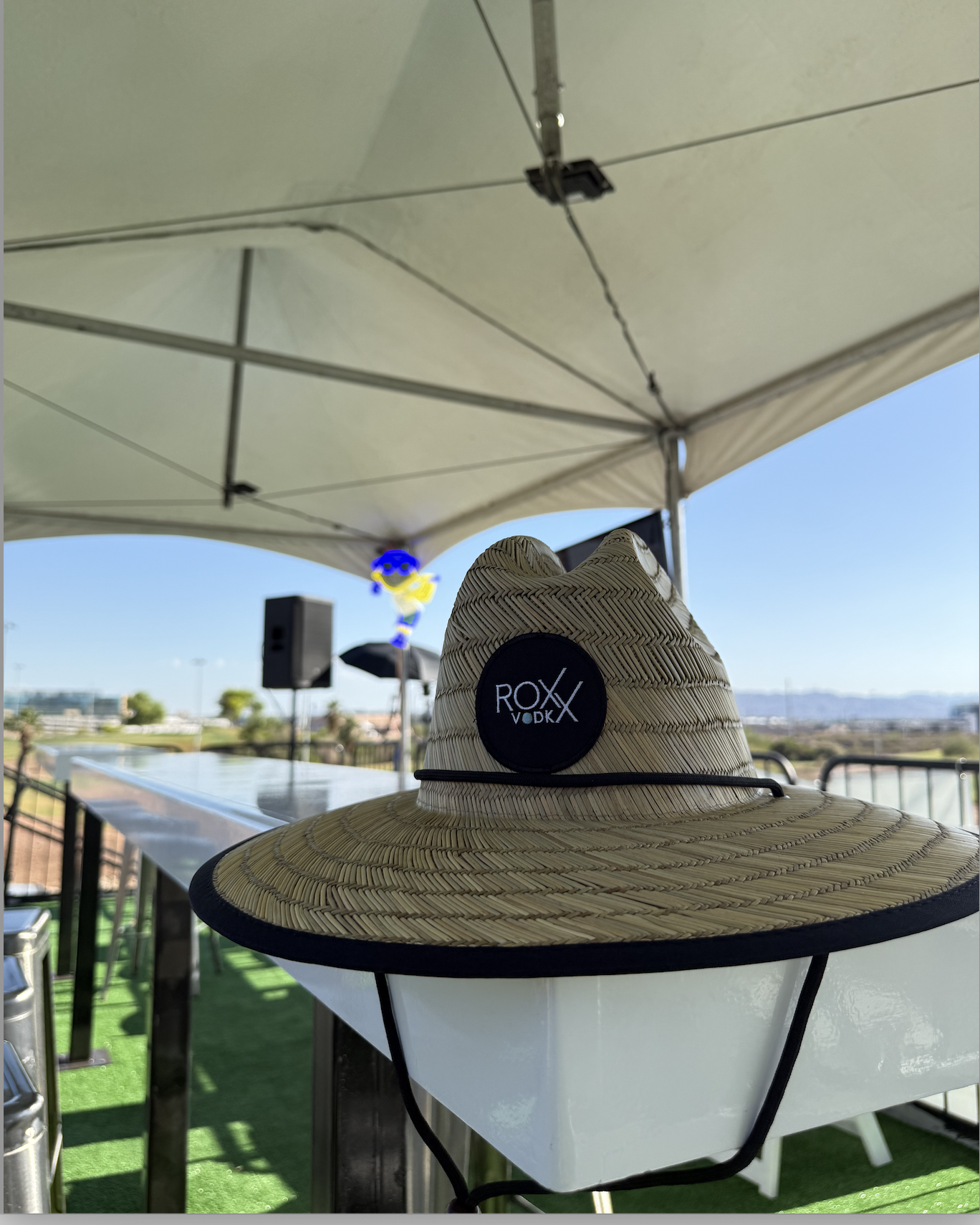 A straw sunhat with a black logo that reads 'ROXX VODKA' on a white table outdoors under a large white patio umbrella, with a blurred background of a blue sky, some clouds, and distant landscape.