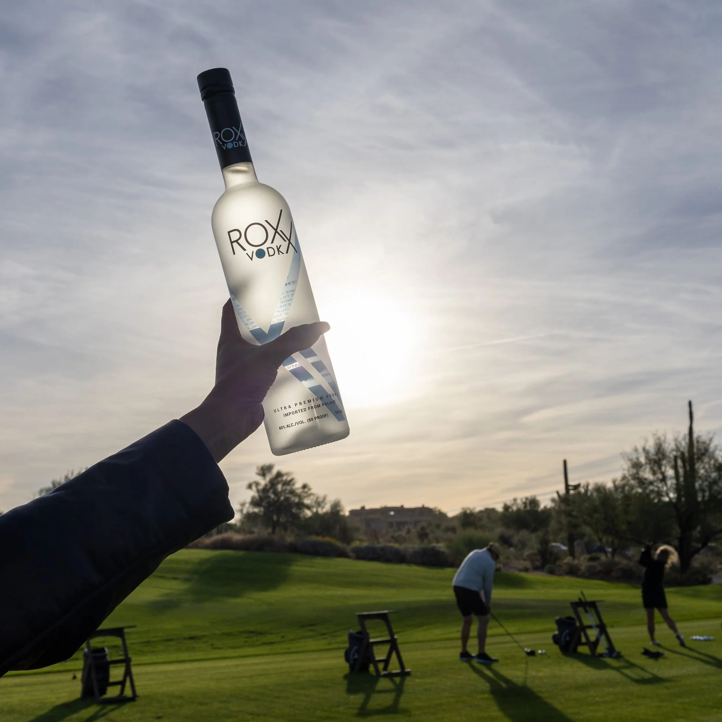 A person holding a bottle of Roxy Vodka against a sunset sky, with three people playing golf on a green golf course in the background.