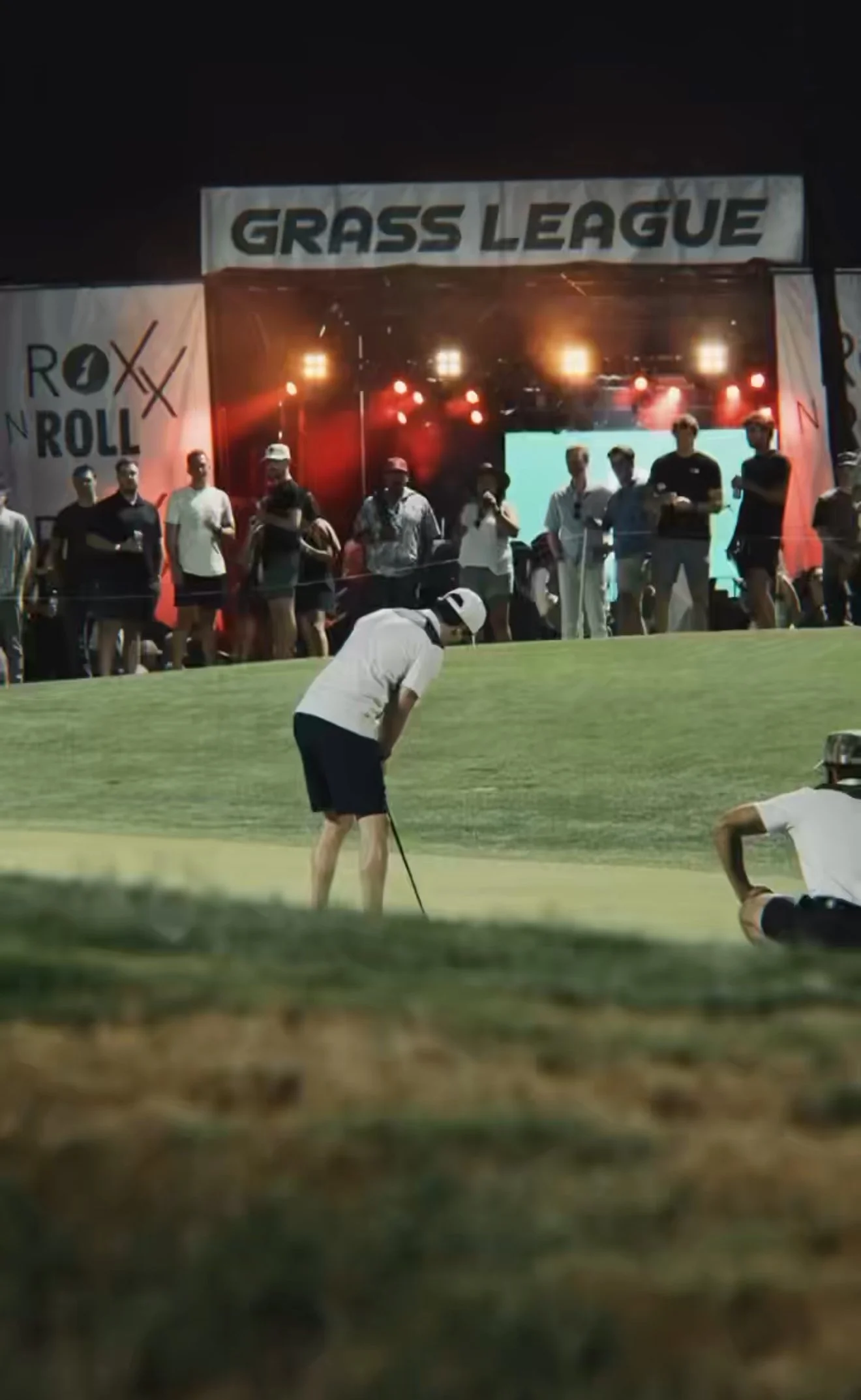 Night scene at a golf tournament with multiple spectators and a player putting on the green, under a stage with a 'Grass League' banner, bright stage lights, and a large screen in the background.