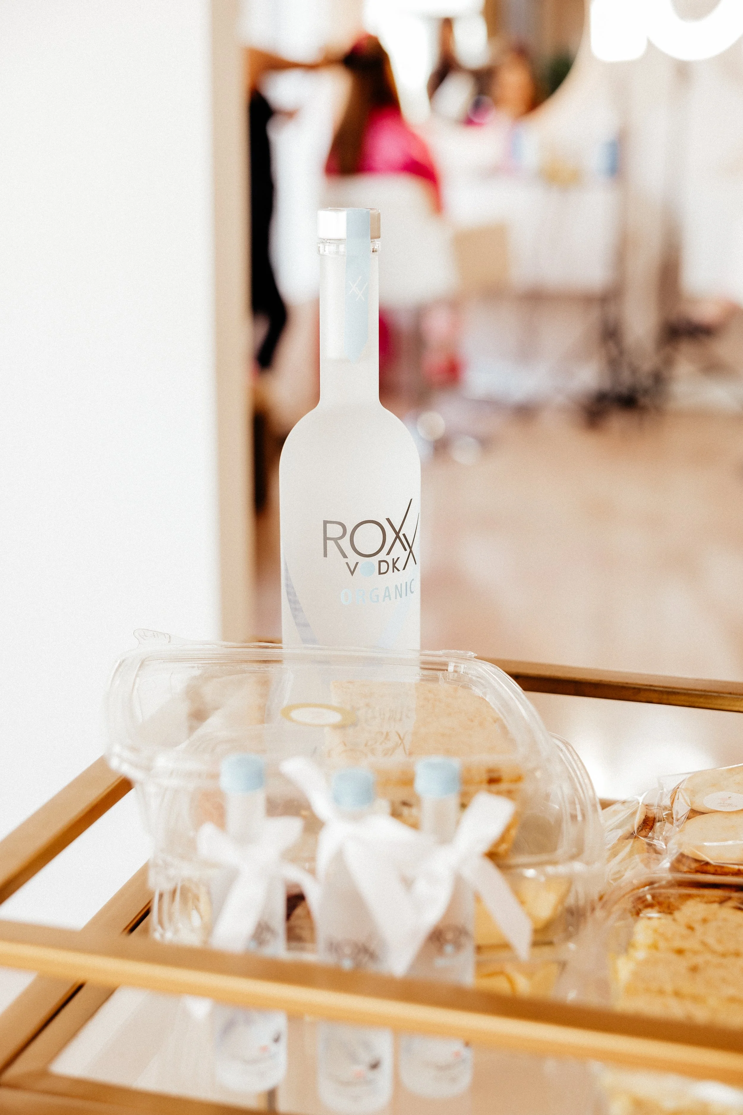 A clear plastic container with bottled beverages labeled 'ROXX VODKA ORGANIC' on a wooden shelf, with a blurred background of people and a bright room.