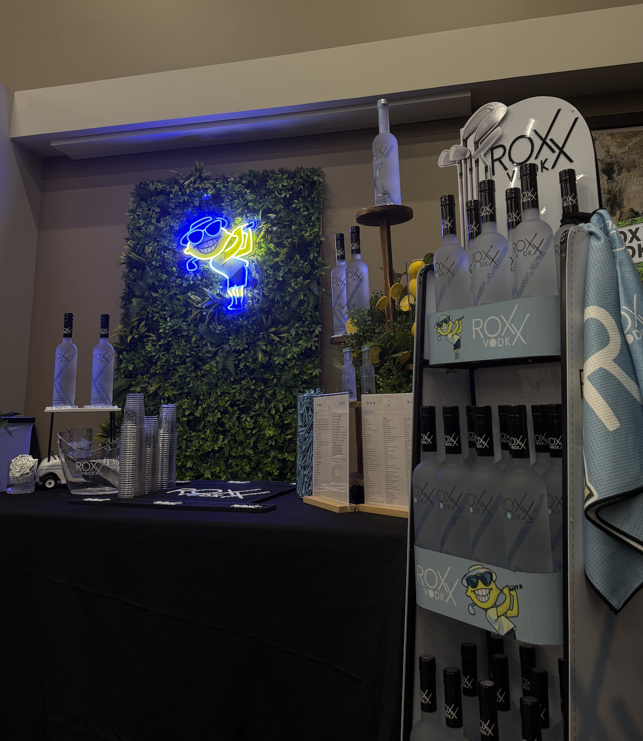 Display of ROX VODK with bottles, neon sign, and decorative greenery at an event or bar setup.