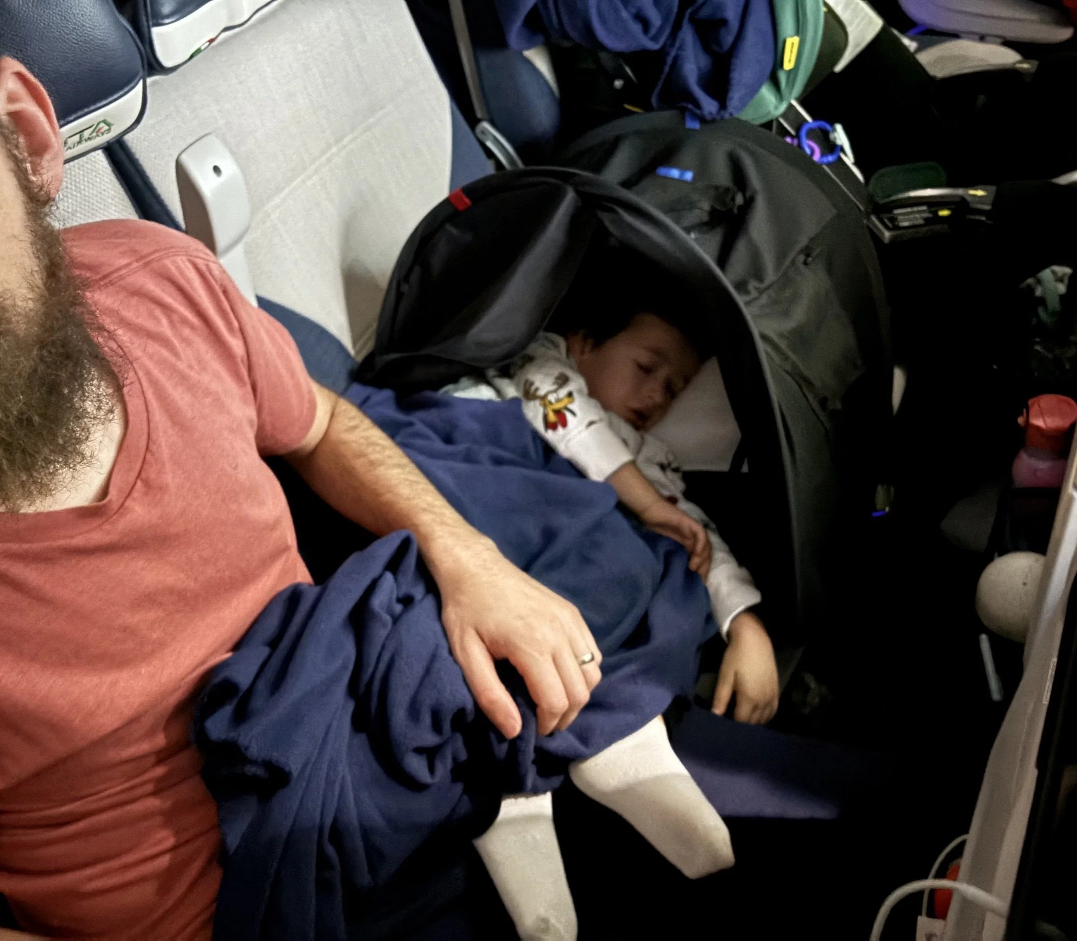 A man with a beard and wearing a salmon-colored shirt is sitting next to a sleeping young boy in a stroller. The boy is resting with eyes closed, wearing a white shirt with colorful designs. Surrounding them are various backpacks and personal items on the seat and in the background.