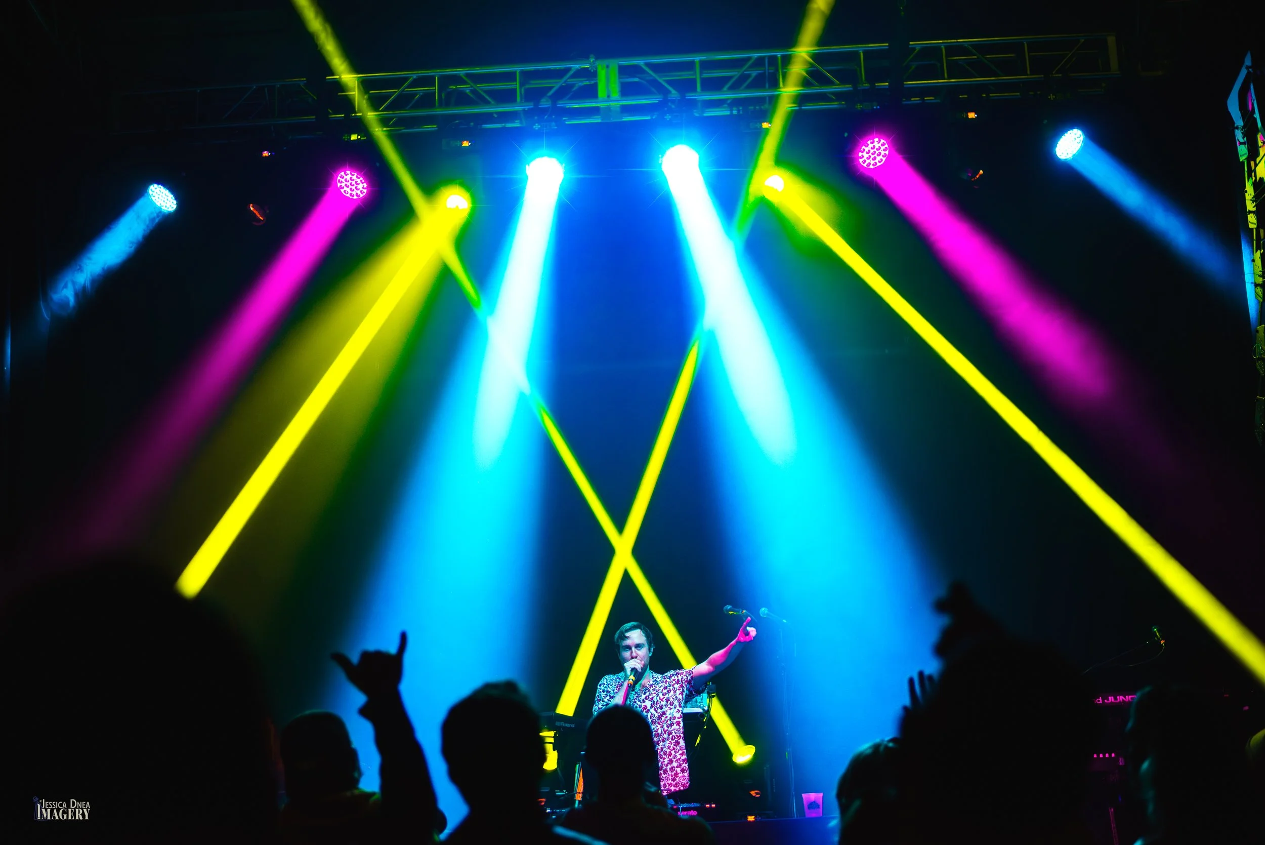 A performer stands beneath intersecting yellow, blue and magenta beams while silhouetted audience members raise their hands at a Denver concert.