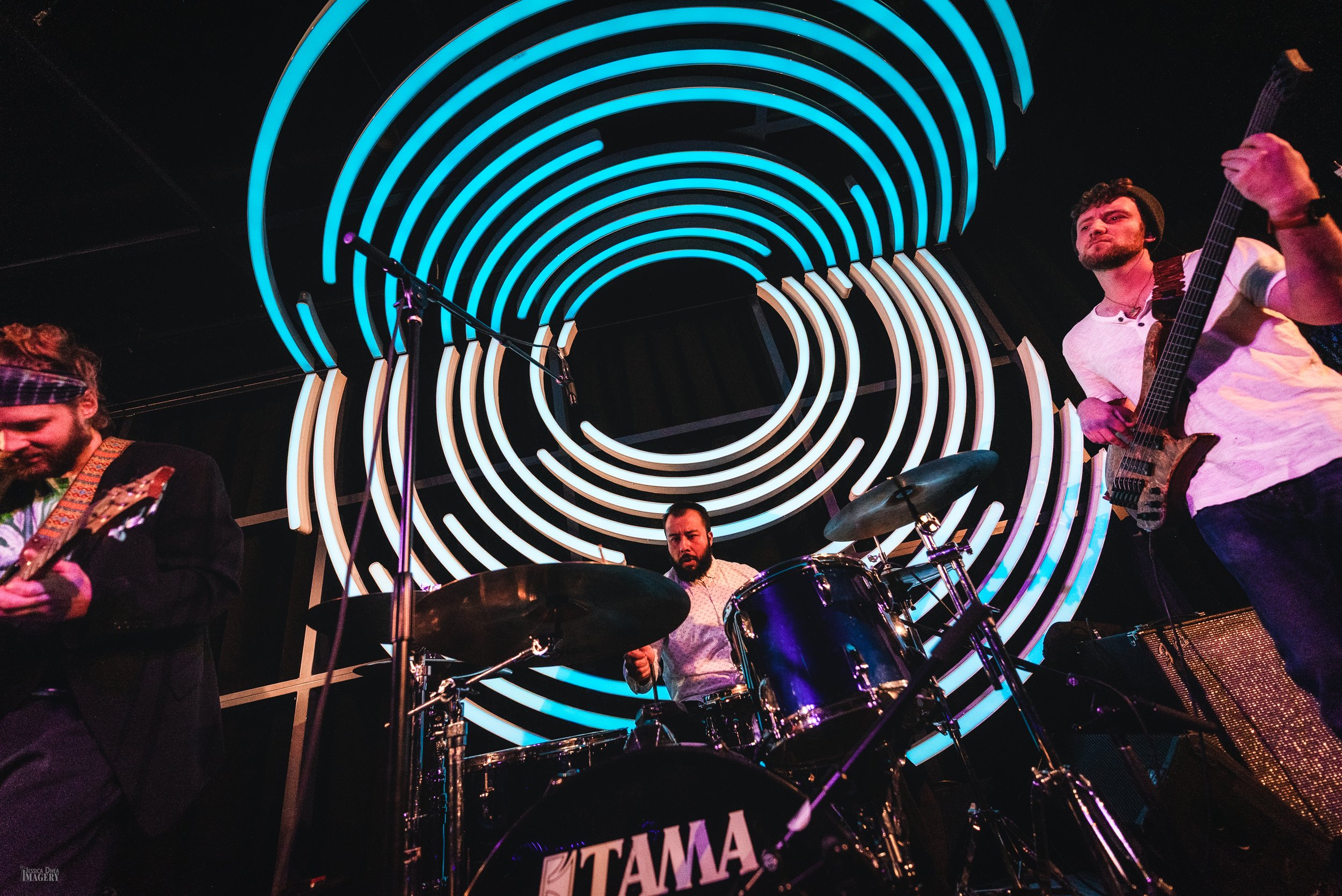 Phooey performs as a trio with the drummer centered inside Dazzle’s repeating blue circular lights during a Denver live-music set.