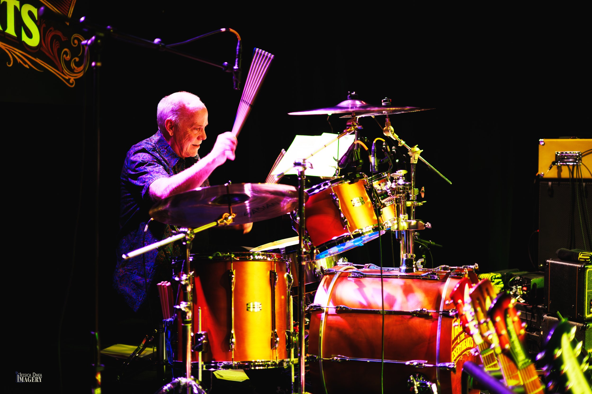 Patrick Dune’s drummer raises both sticks over an orange drum kit during an energetic rock concert photographed in Denver.