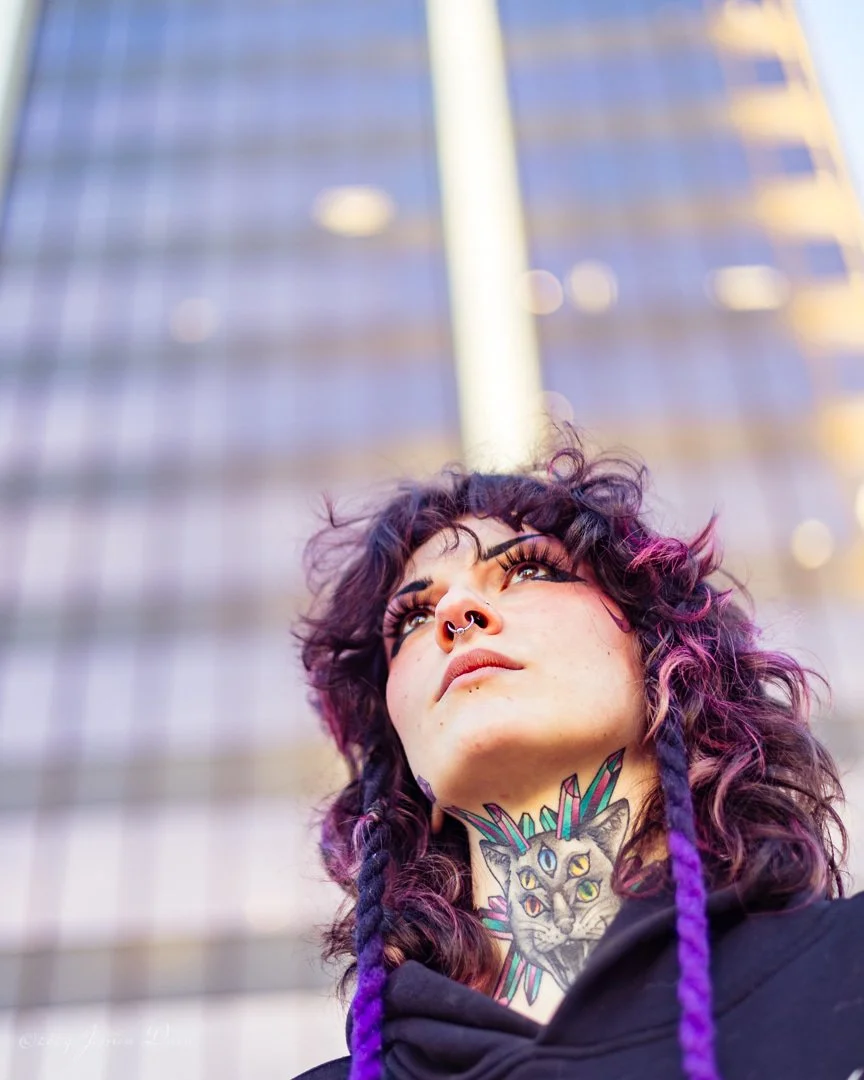 Low-angle personal branding portrait of a tattoo artist with purple hair and visible neck art against reflective downtown architecture.

