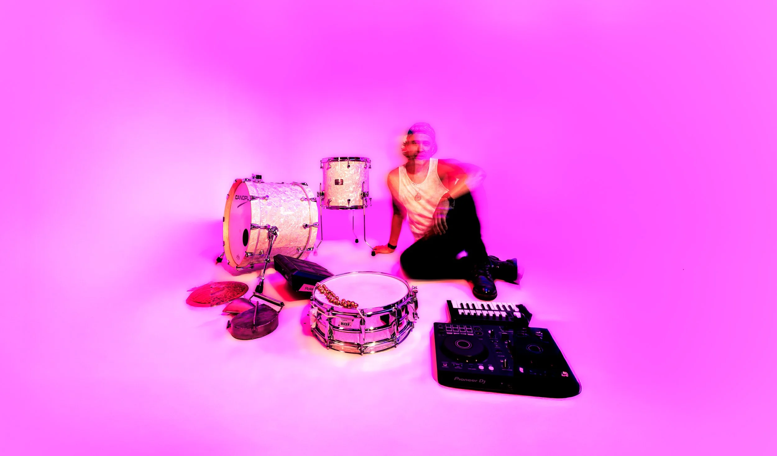Creative artist press photo with drums, DJ equipment and vivid pink studio lighting on a white cyc wall.
