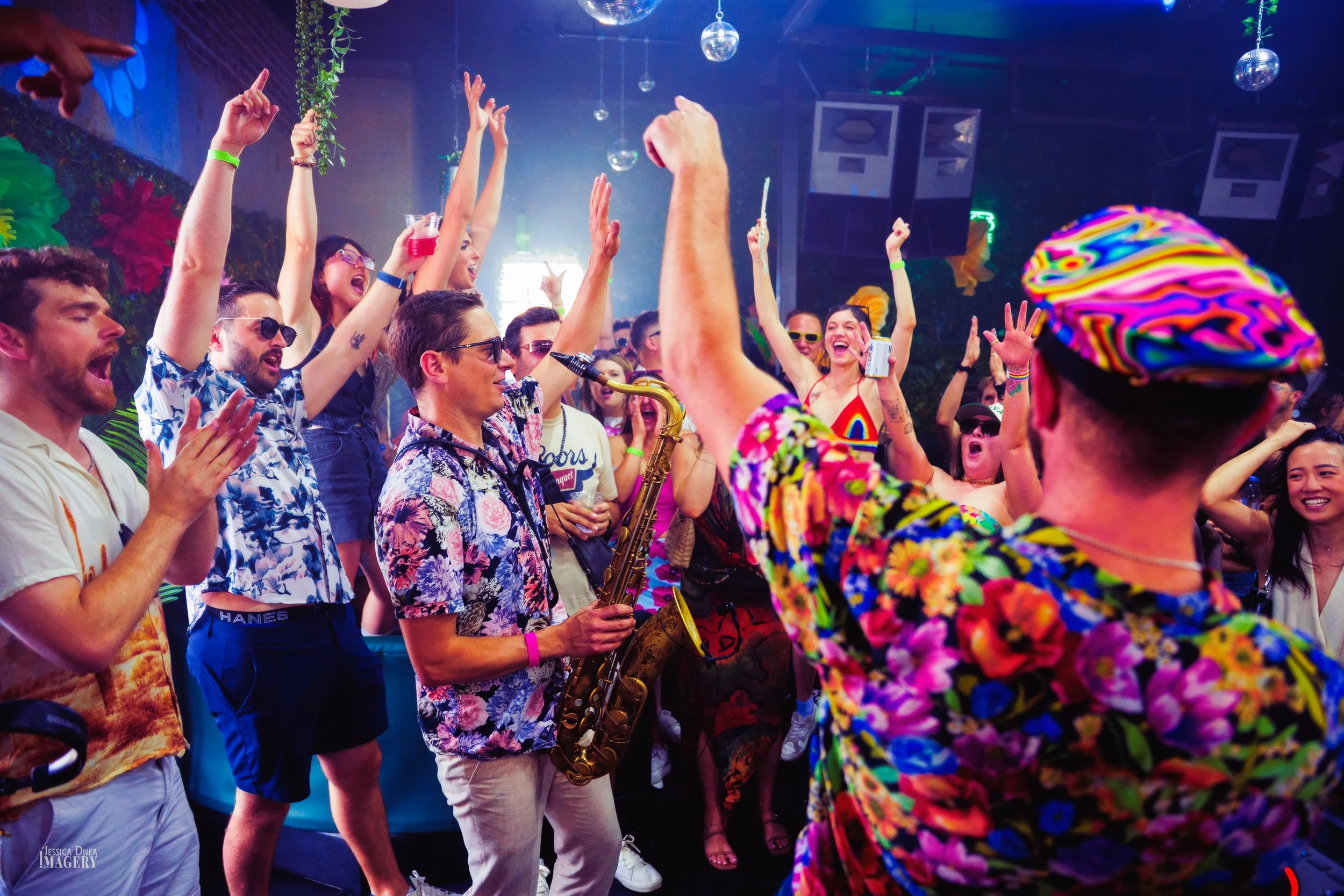 A saxophonist performs in the center of an enthusiastic Pride crowd with raised hands during a colorful Denver music event.