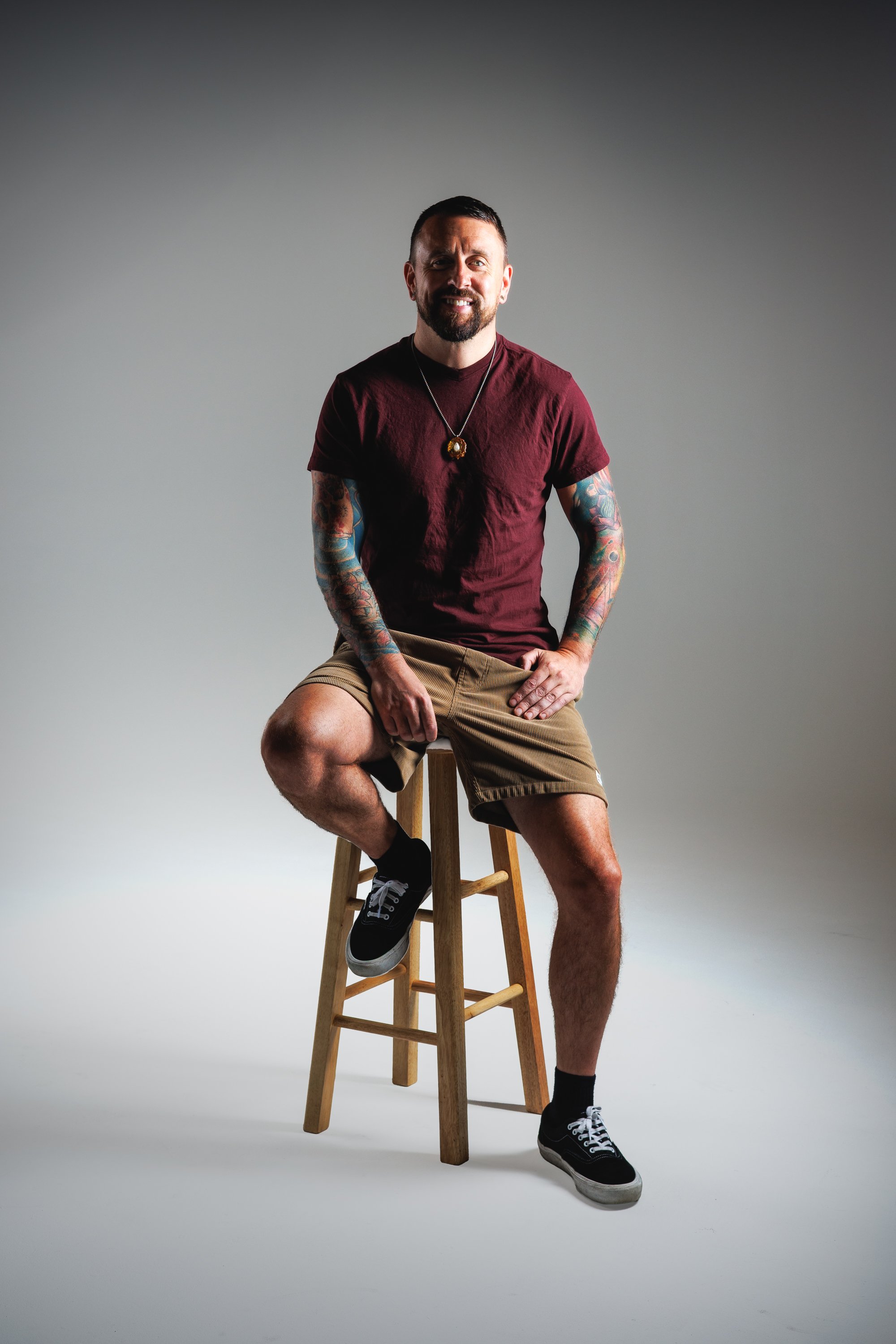 Full-length studio branding portrait of a tattooed man smiling on a wooden stool by Denver photographer Jessica Dnea Imagery.