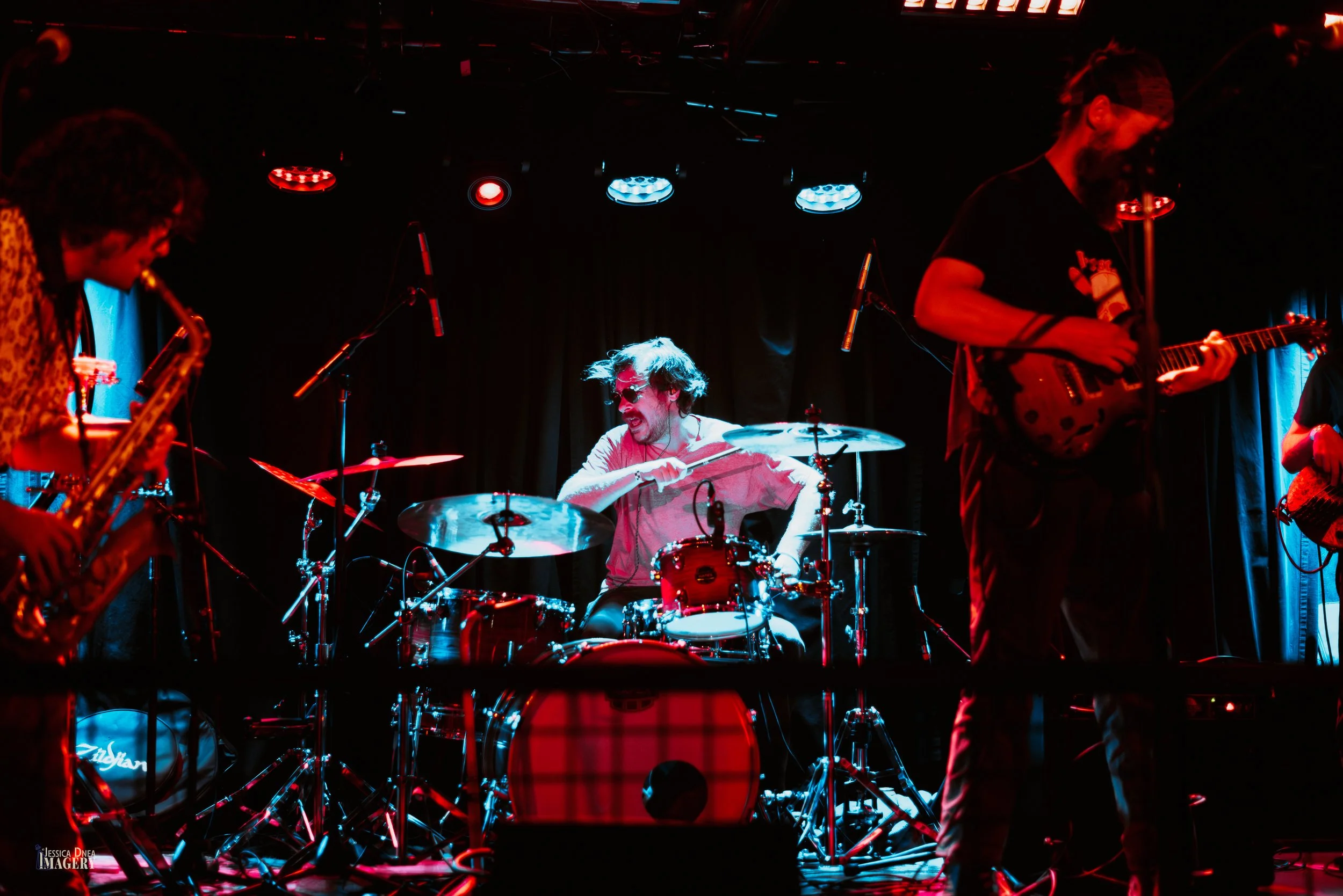 Phooey’s drummer performs beneath red stage lighting while guitarists frame the kit during a live Denver band concert.