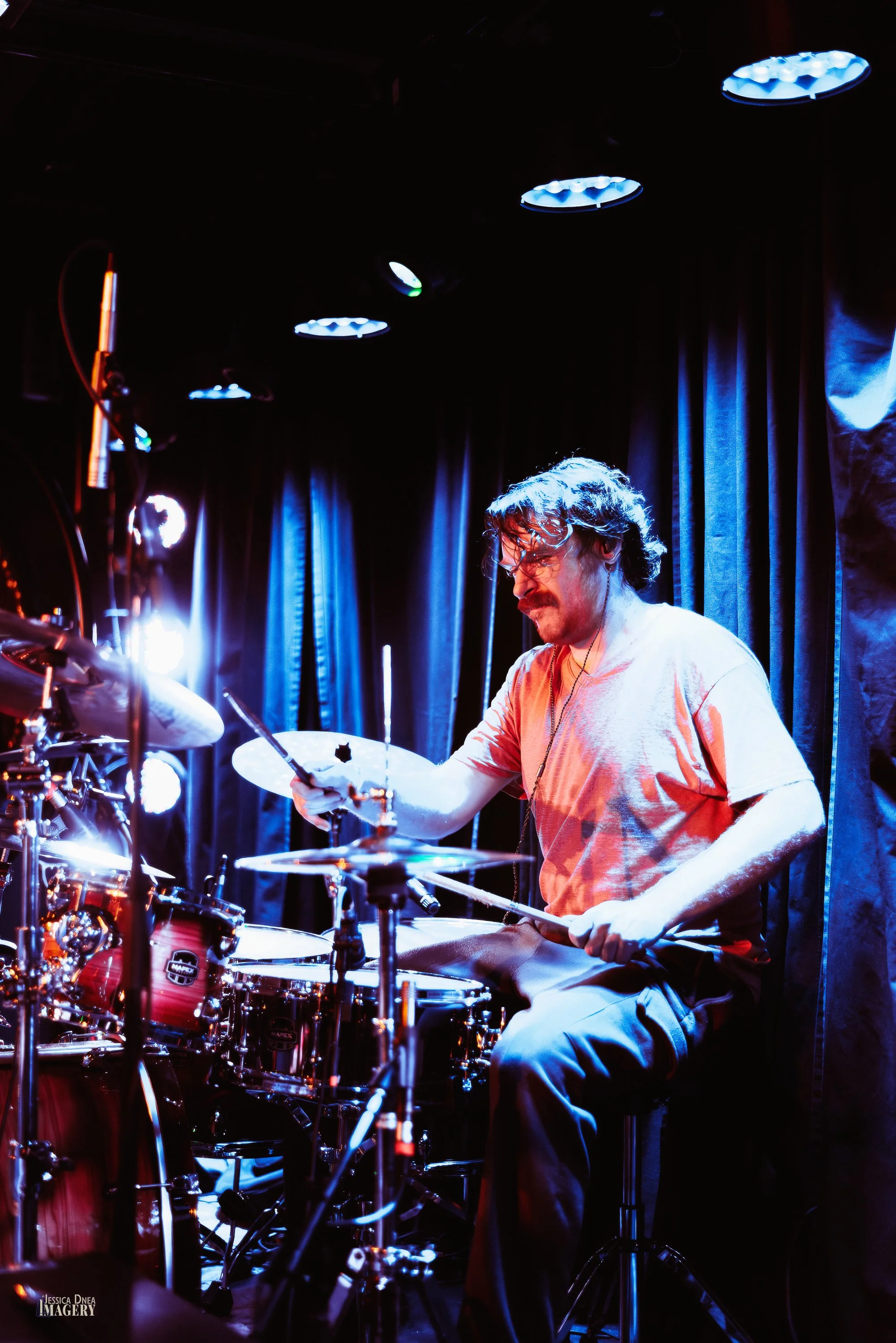 Phooey’s drummer plays beneath blue, white and red stage lights in a vertical Denver concert photograph by Jessica Dnea.