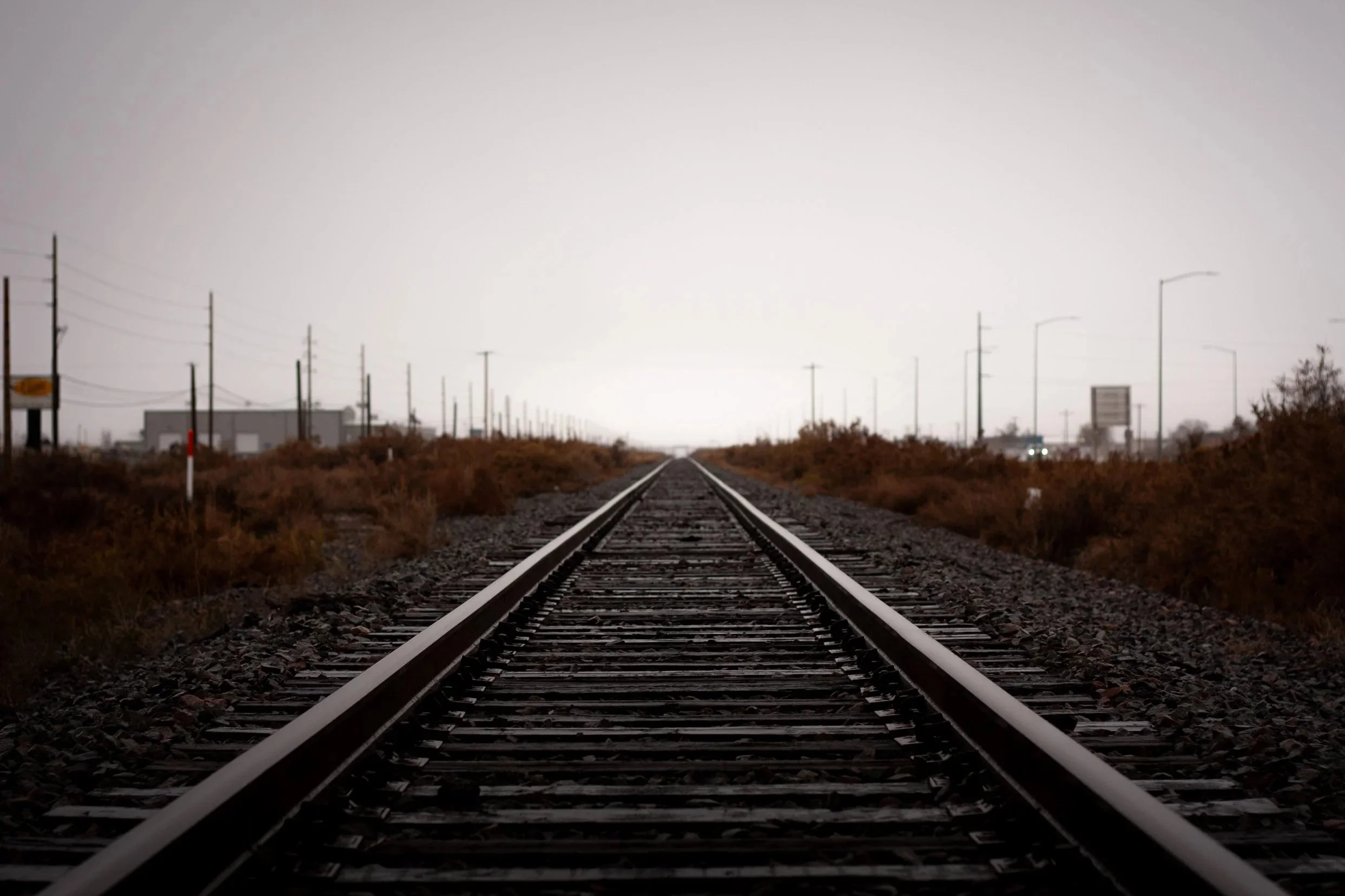 A straight railway track extending into the distance with overcast sky, surrounded by dry bushes and utility poles lined along the sides.