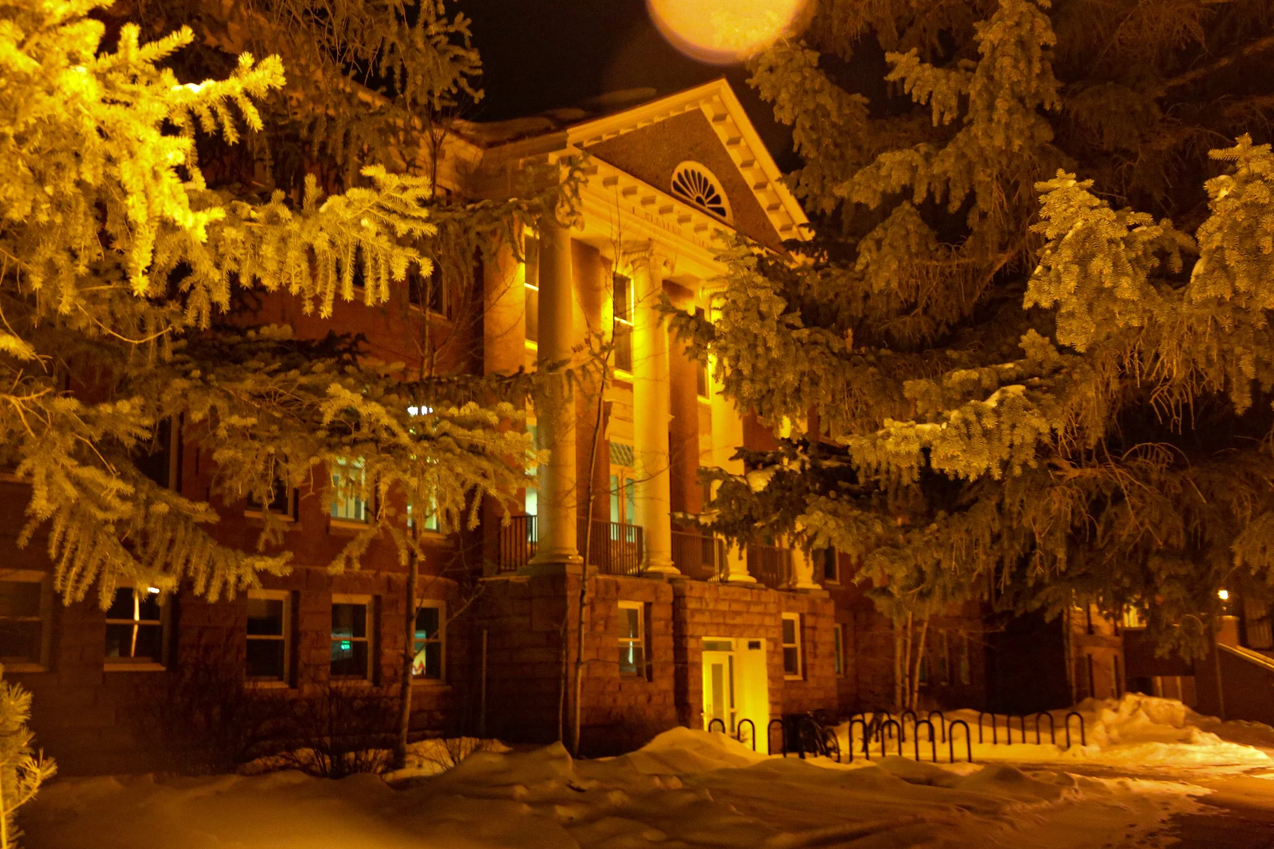 Historic building illuminated at night, surrounded by snow-covered trees, with columns and warm lighting.