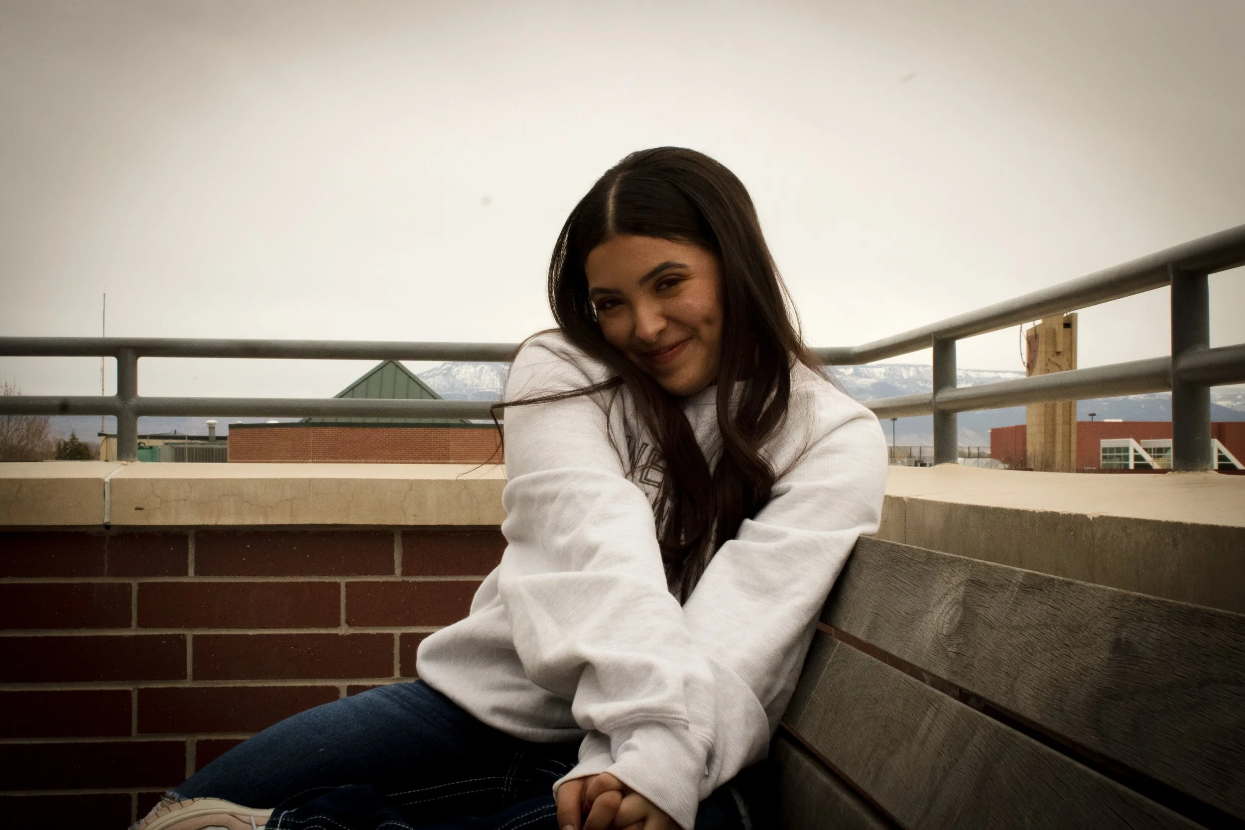 Person sitting on a rooftop bench, smiling, wearing a white sweatshirt, with a cloudy sky and buildings in the background.