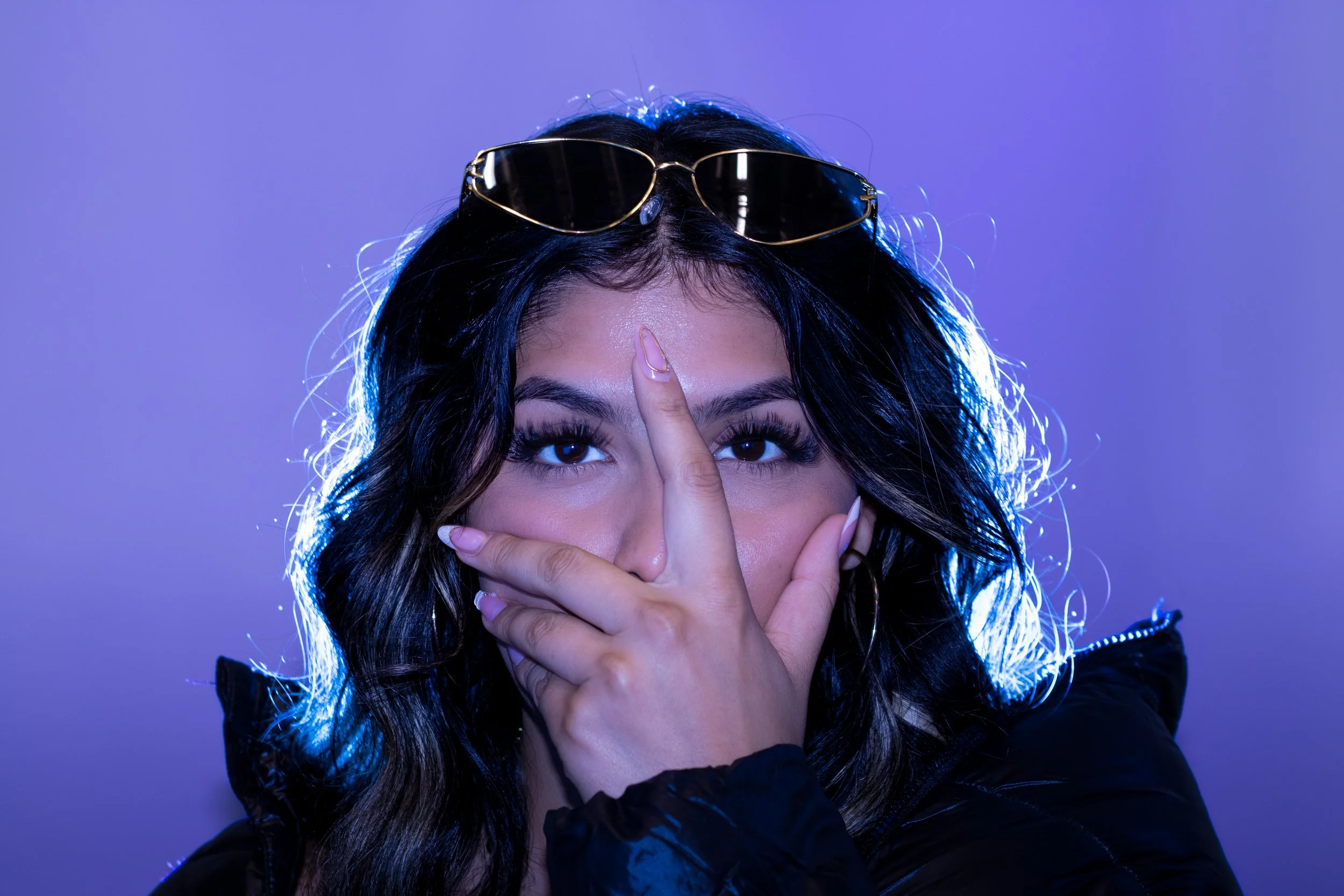 A woman with dark hair, wearing sunglasses on her head, holding her hand in front of her face in a 'peace' sign, against a purple background.