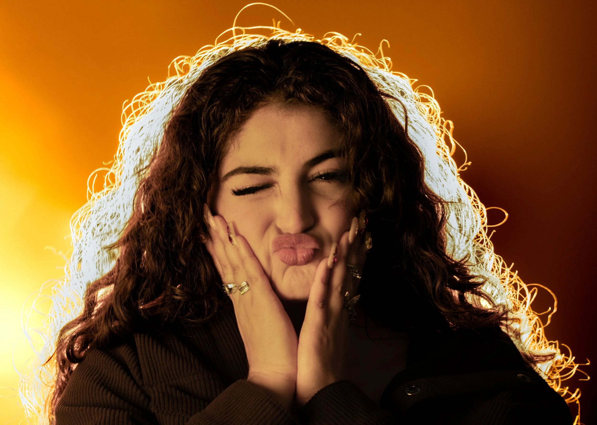 A woman with curly hair making a playful, puckered face, holding her cheeks with her hands, against a warm yellow-orange background.