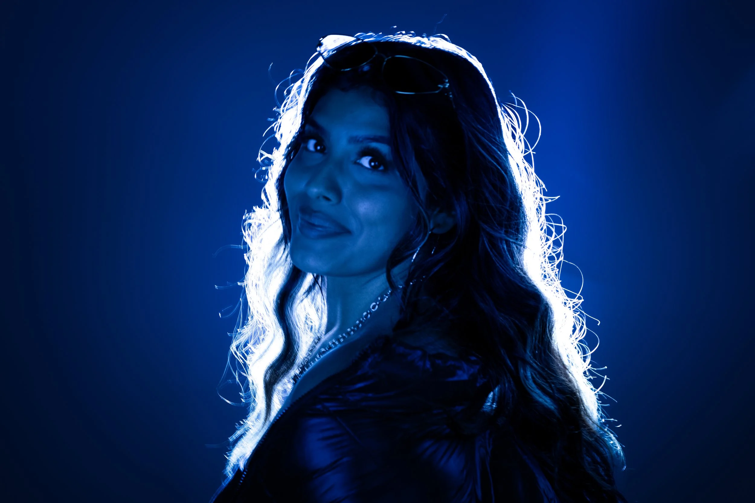 A woman with long wavy hair, wearing sunglasses on her head and a leather jacket, is illuminated by blue backlighting, creating a dramatic silhouette.