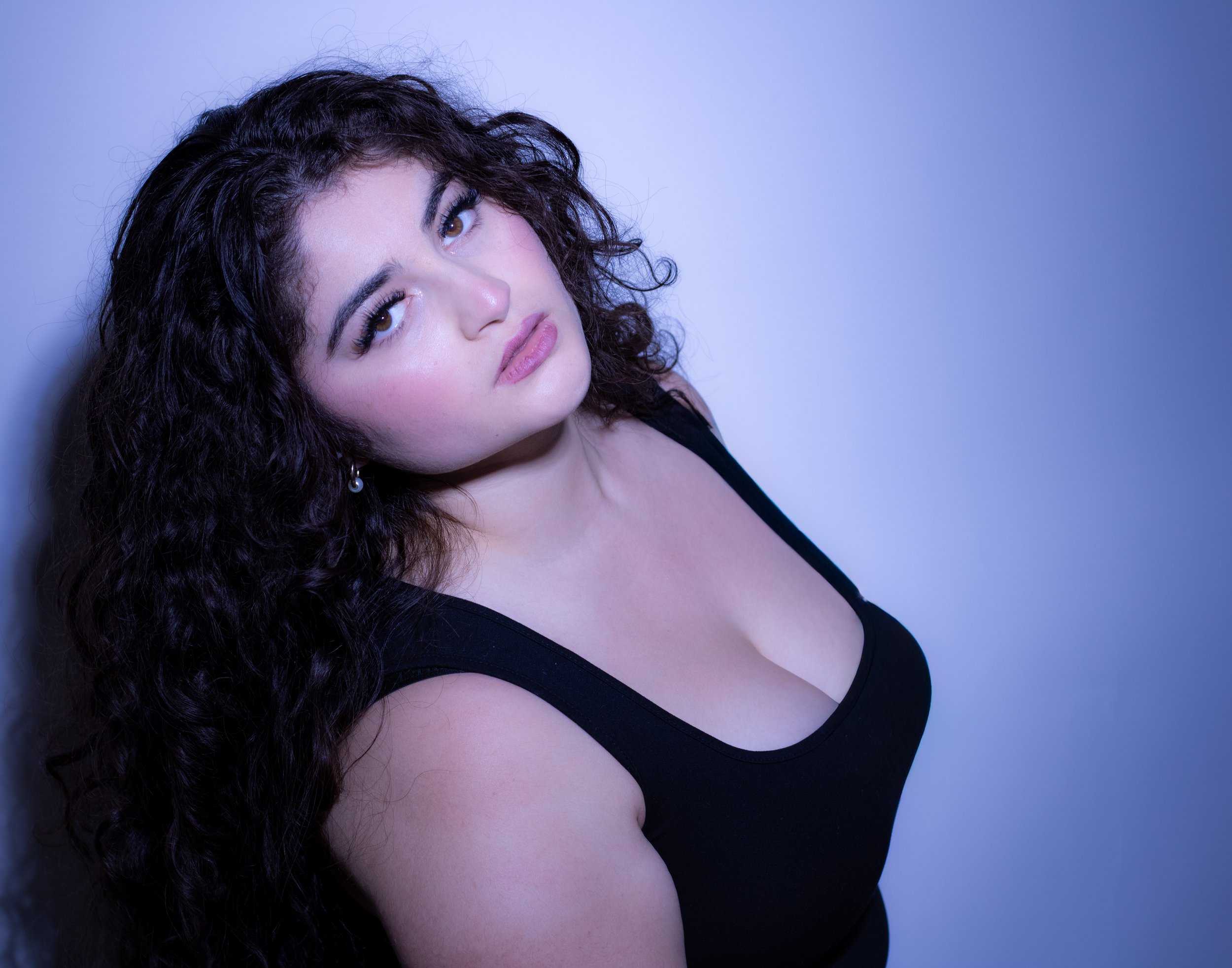 A young woman with long, curly dark hair and light skin leaning against a wall, wearing a black tank top, looking at the camera with a neutral expression, with bluish-purple lighting.