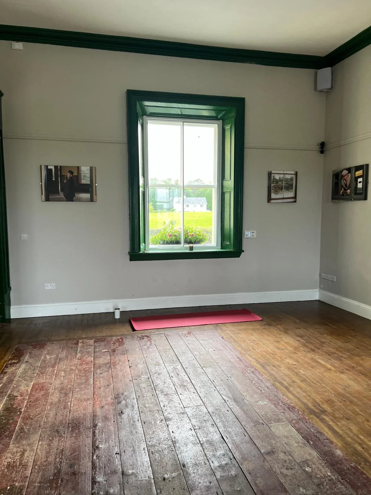 Empty room with hardwood floor, open window with green shutters, flower box outside, three photographs on walls, electrical outlets, and a pink yoga mat on the floor.