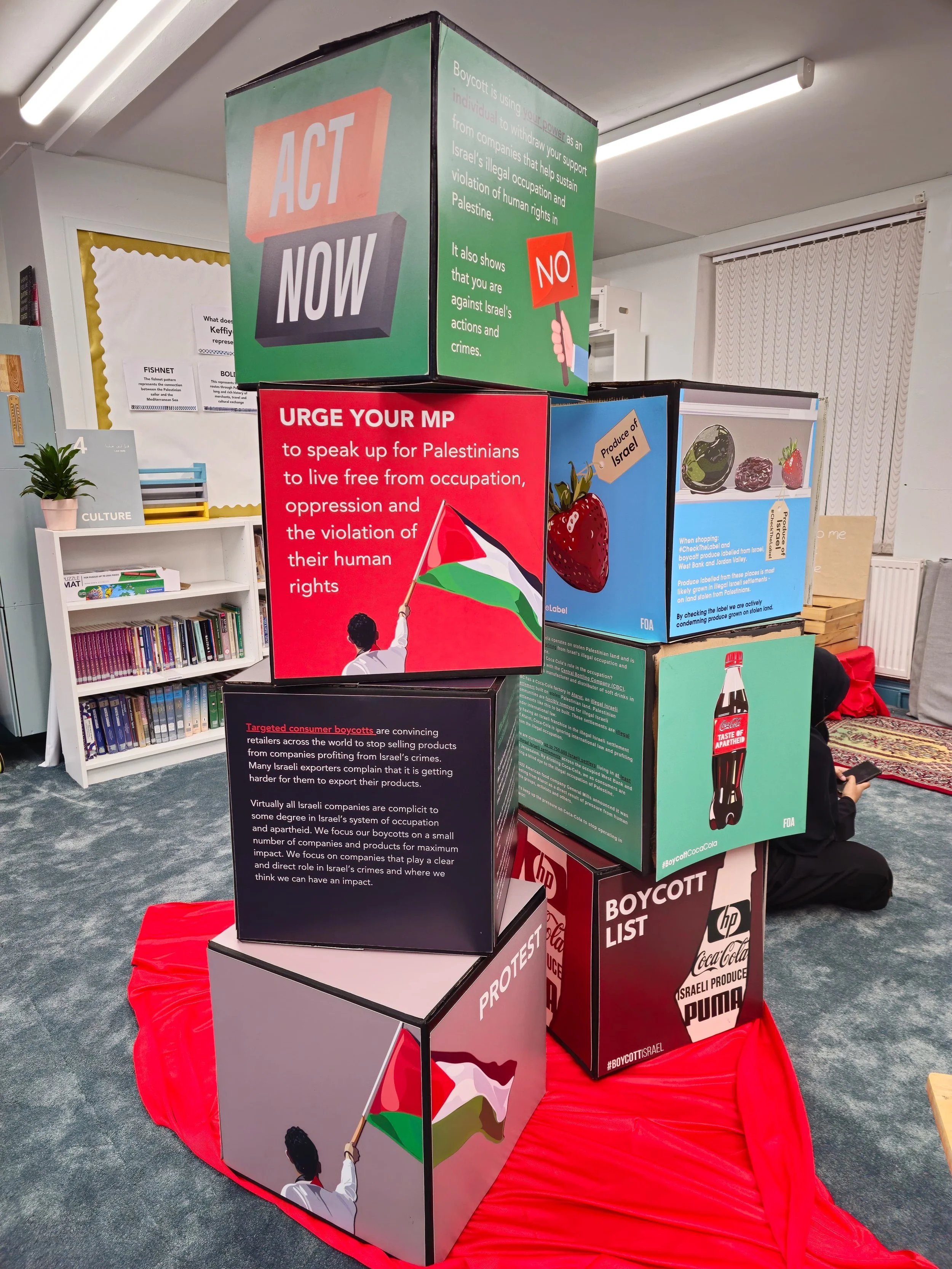 Colorful display cubes with pro-Palestinian messages and images, including flags, strawberries, a soda bottle, and boycott-related posters, set on a red fabric at an indoor location.
