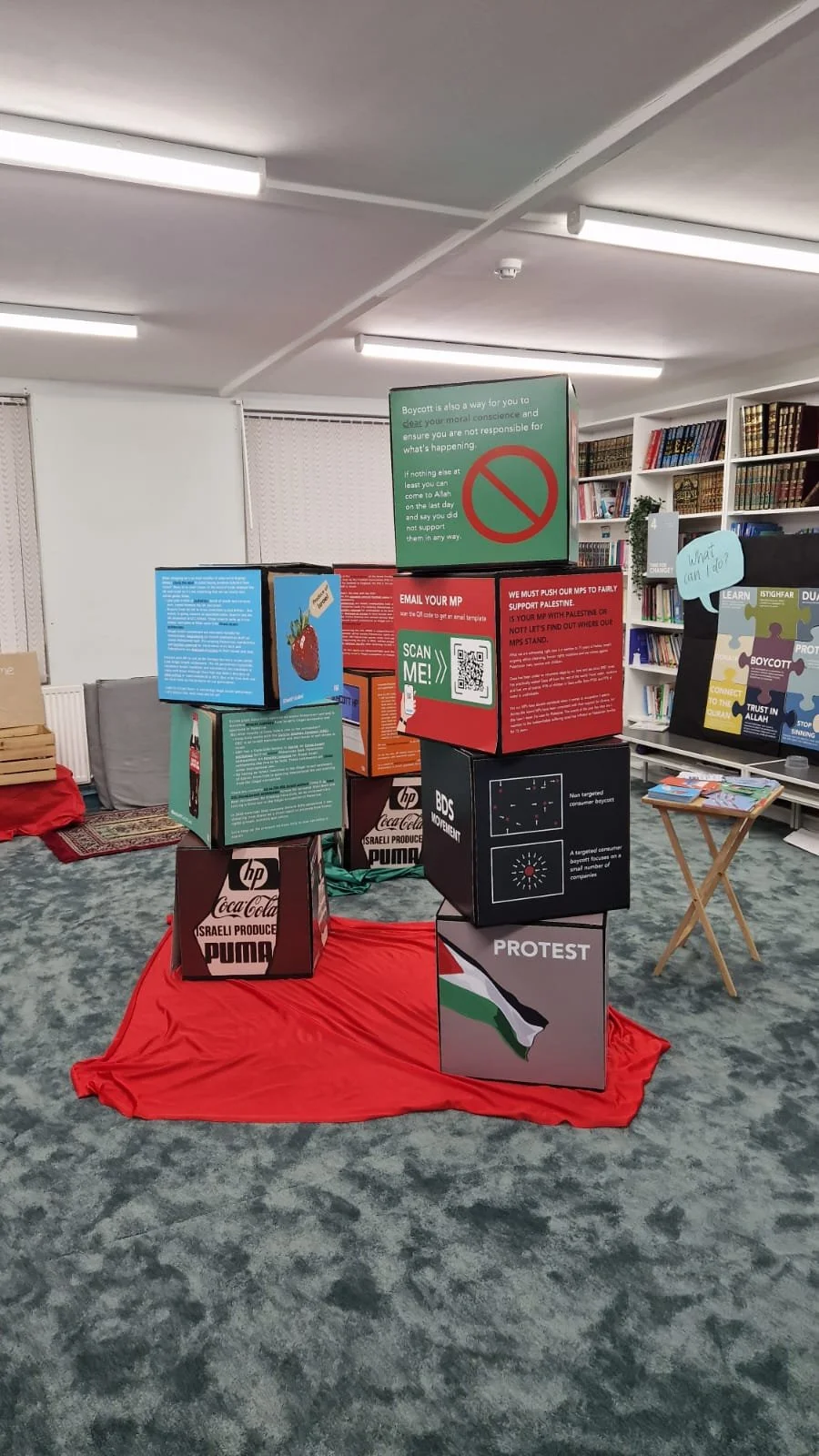 Display of boxes with various messages and symbols on a red cloth on the floor in a room with bookshelves and a table with books.