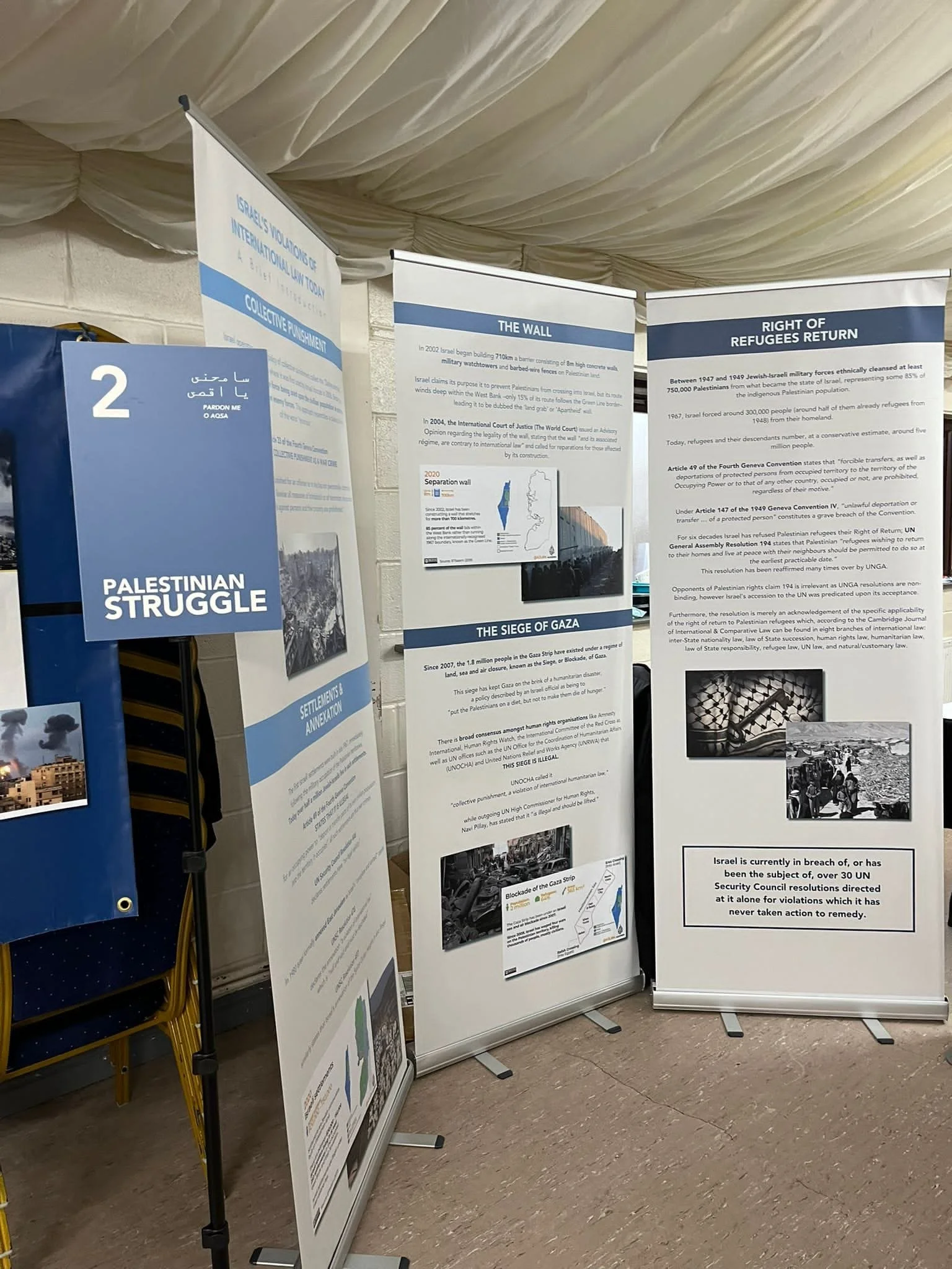 Three informational display boards with text and images about Palestine, Gaza, and Israeli policies, set up in an indoor location with chairs on the side.
