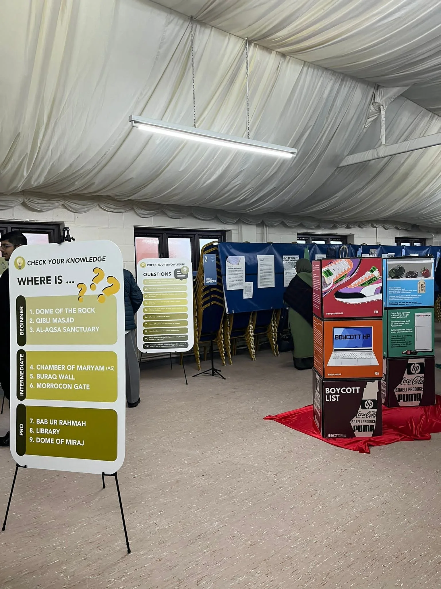 Indoor event with informational display boards and a red cloth covering a platform that holds election boycott posters, including logos of Coca-Cola, HP, and Puma, with a person wearing a green headscarf and dark jacket in the background.