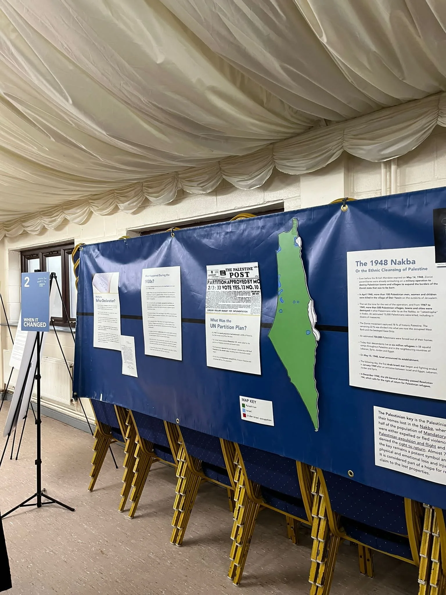 Historical display with information board about the 1948 Nakba, featuring a map of Palestine marked in green and orange, and papers with text mounted on a blue background. A sign on a stand reads "When It Changed."