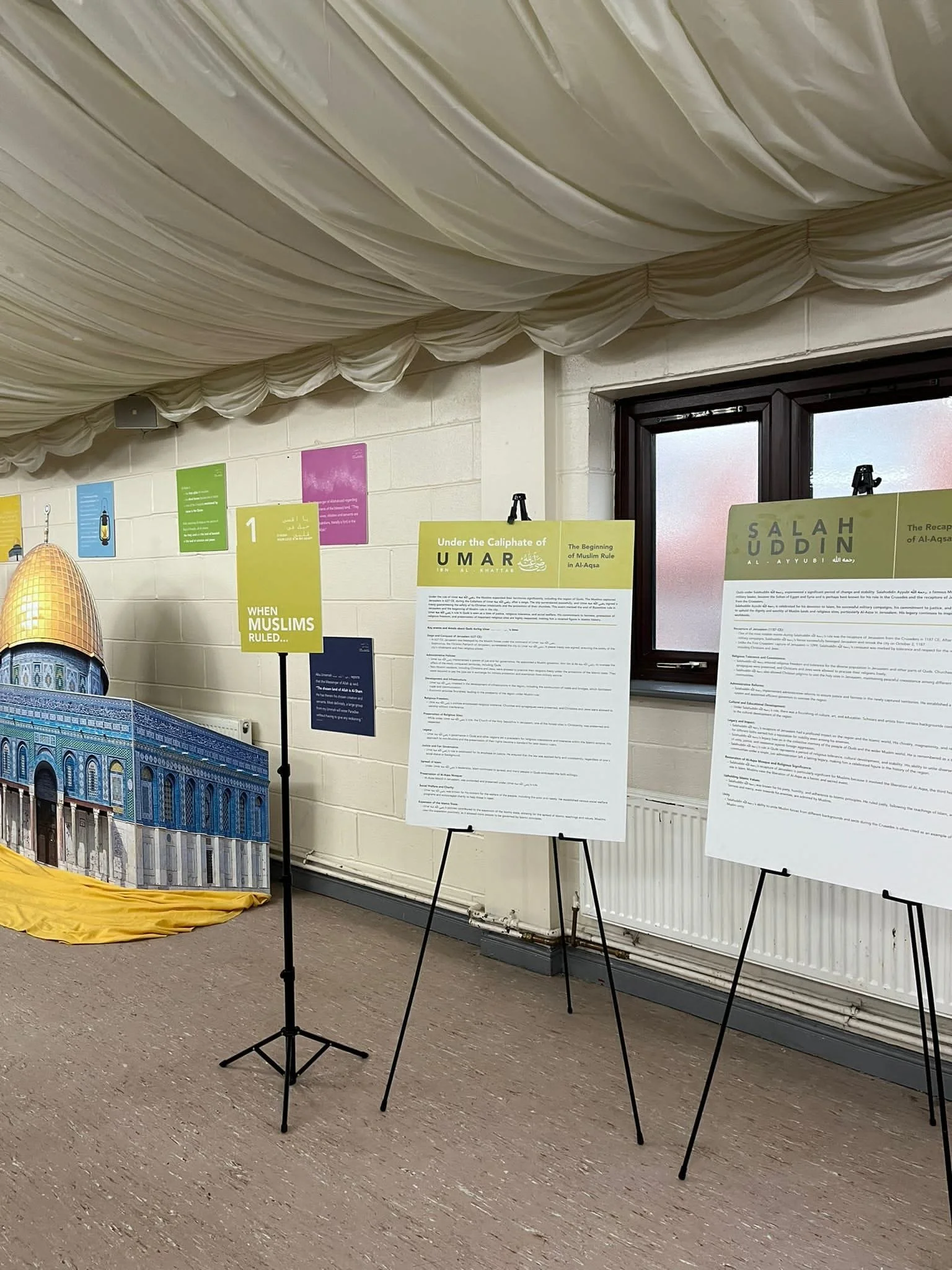 Exhibition display with informational posters on easels, depicting historical and religious topics, set inside a room with cream-colored bricks, a window, and decorative fabric ceiling draping.