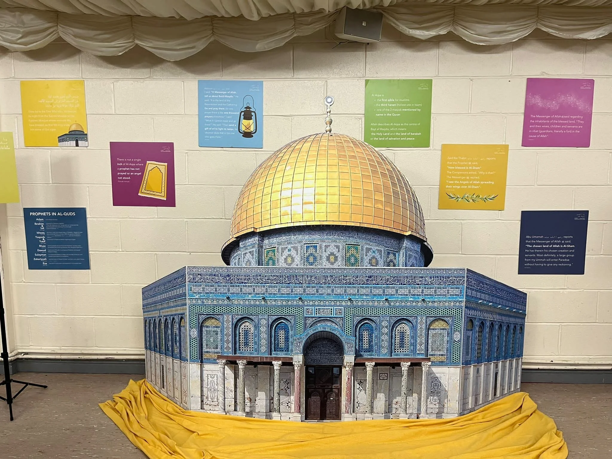 A detailed model of the Dome of the Rock, an Islamic shrine, placed on a yellow cloth. The model showcases the shrine's intricate blue tilework and gold dome. Behind it, a wall with colorful informational posters about prophets in Islam.