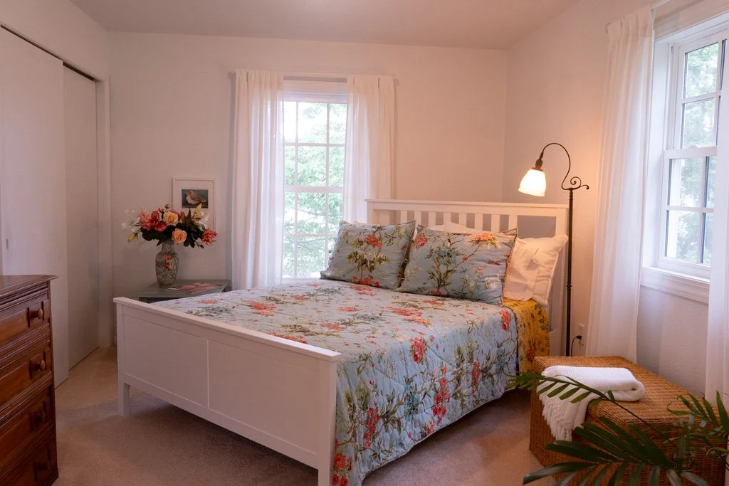 A cozy bedroom with a white bed frame, floral bedspread, and matching pillows. There are two windows with white curtains, a bouquet of flowers on a bedside table, and a wicker basket with towels and a plant in the corner.