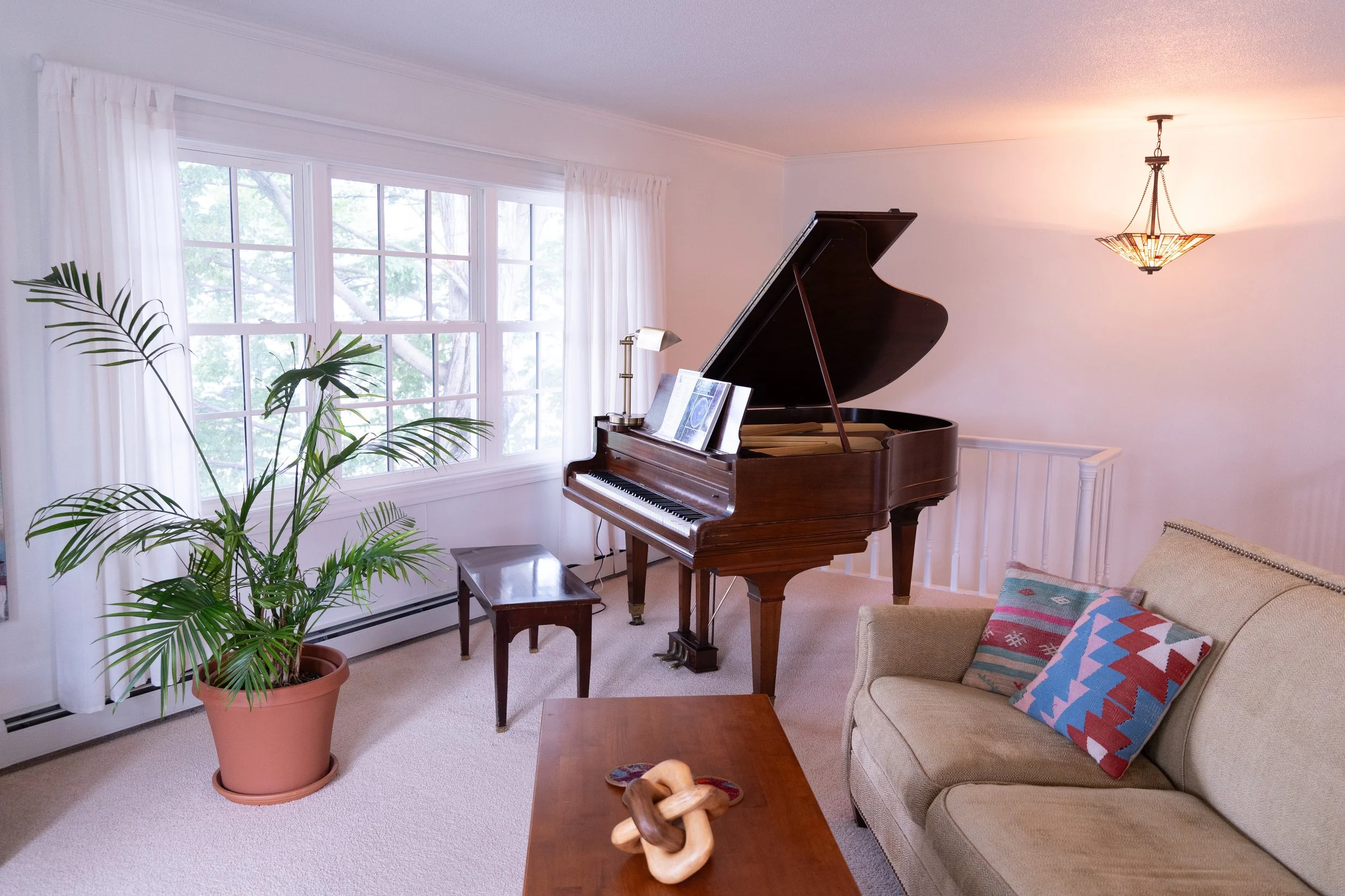 Living room with grand piano, potted plant, window with white curtains, beige sofa with colorful cushions, wooden coffee table with wooden knot decor, small bench, and ceiling light fixture.
